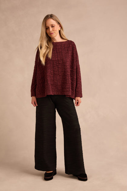 A woman wearing the Bordeaux Textured Squares Jersey Boxy Top and loose black pants stands in front of a neutral beige background.