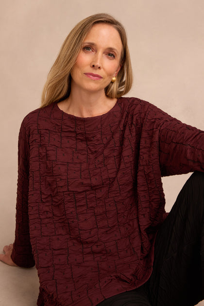 A woman with long blonde hair sits against a plain beige background, wearing the Bordeaux Textured Squares Jersey Boxy Top and looking at the camera with a relaxed expression.
