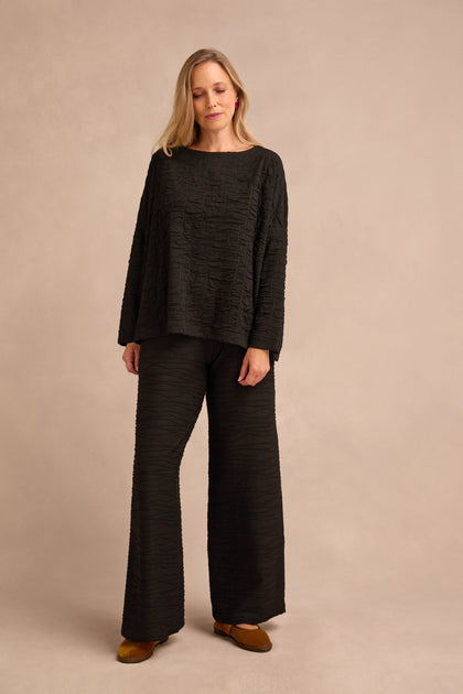 Woman standing against a beige background, wearing the Black Textured Squares Jersey Boxy Top with matching wide-leg pants and brown shoes.