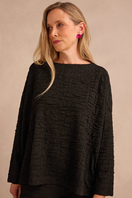 A woman with long blonde hair is wearing the Black Textured Squares Jersey Boxy Top and bright pink earrings, standing against a neutral background.