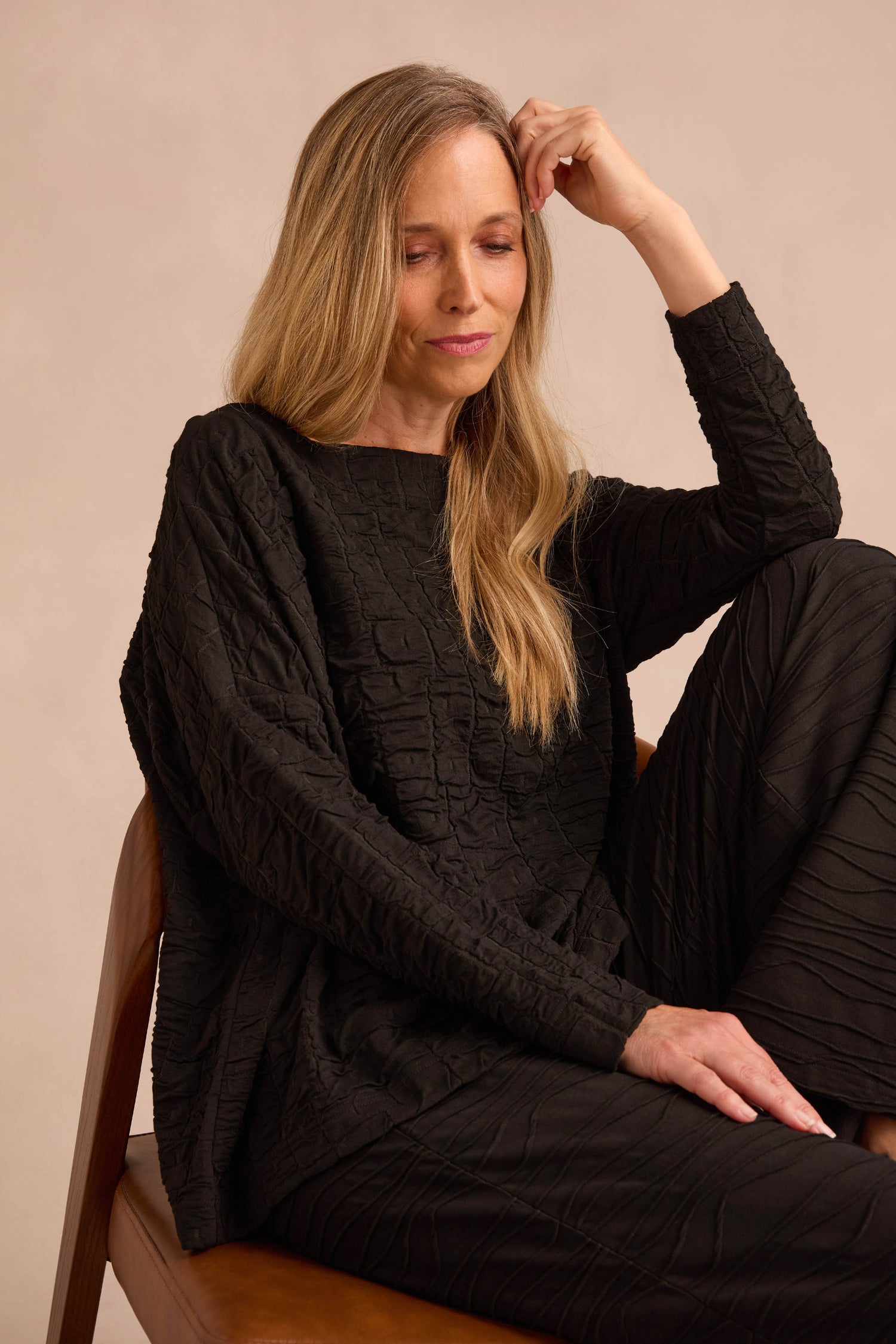 A woman with long blonde hair sits on a chair wearing the Black Textured Squares Jersey Boxy Top and matching pants, resting her arm on her knee and looking down.