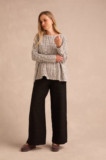 A woman stands against a beige background, wearing the Silver Textured Squares Jersey Boxy Top paired with wide-leg black pants and mauve shoes, one hand touching her hair while the other rests on her arm.