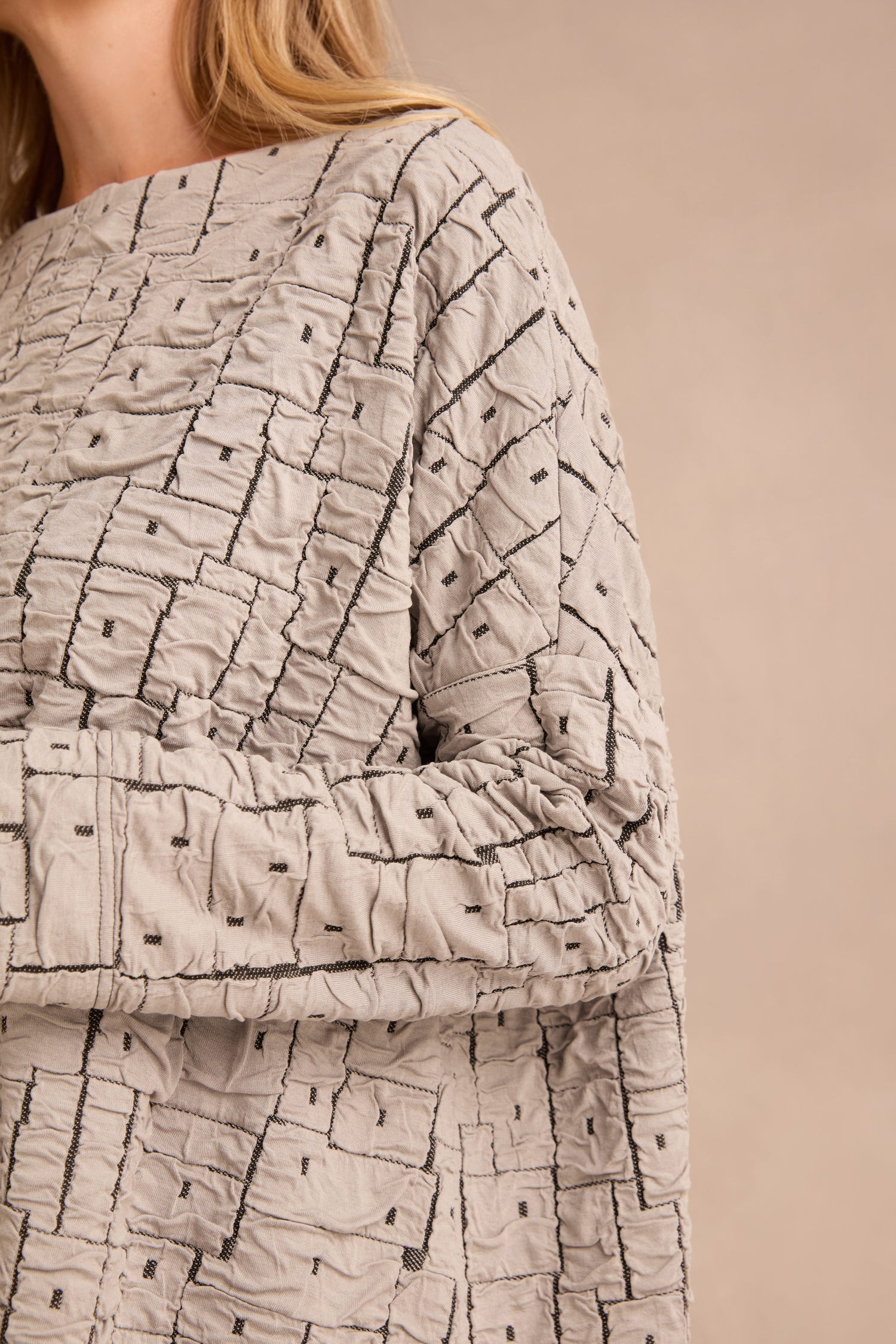 A person is wearing the Silver Textured Squares Jersey Boxy Top, a light gray, long-sleeve boxy top featuring a geometric stitched pattern; only their upper body and part of their face are visible.