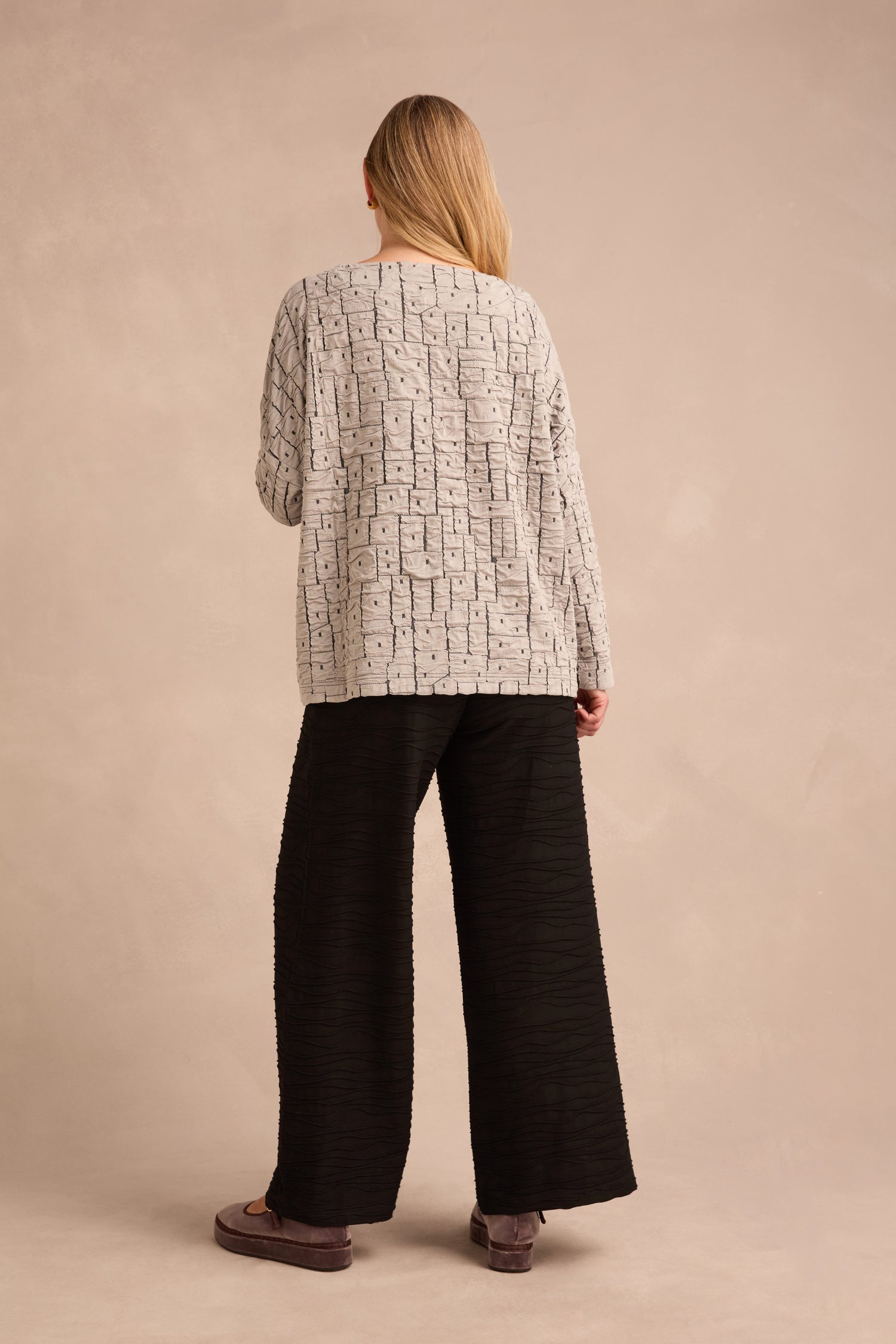 A person with long hair stands facing away, wearing the Silver Textured Squares Jersey Boxy Top, wide black pants, and brown shoes against a neutral background.