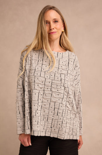 A woman with long blonde hair is wearing the Silver Textured Squares Jersey Boxy Top in light gray, styled with black pants and standing against a plain beige background.