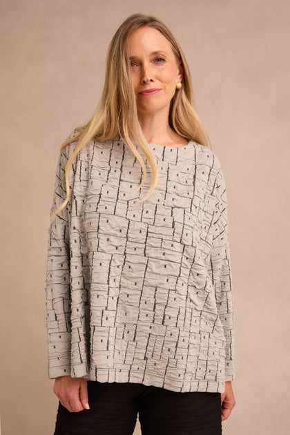 A woman with long blonde hair is wearing the Silver Textured Squares Jersey Boxy Top in light gray, styled with black pants and standing against a plain beige background.
