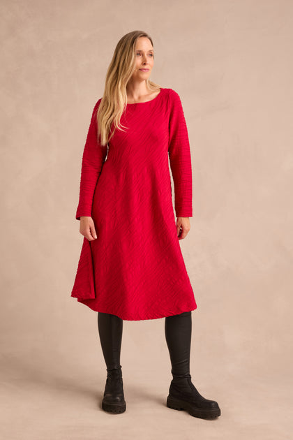 A woman stands against a plain beige background, wearing the Wave Textured Jersey Flared Dress in red—a long-sleeve, knee-length flared design—paired with black leggings and black shoes. Her long blonde hair is draped over one shoulder.