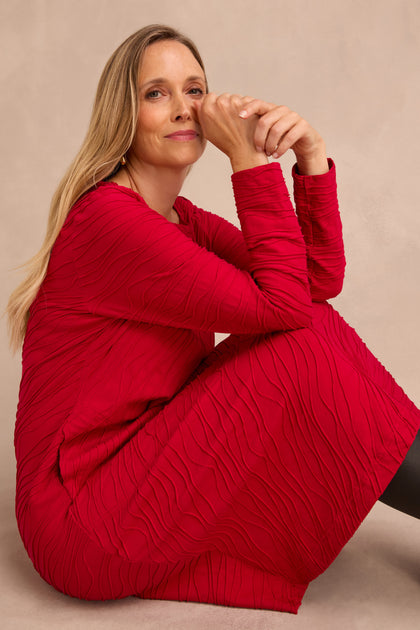 A woman with light hair sits on the floor, looking at the camera with a relaxed expression, wearing the Wave Textured Jersey Flared Dress in red with long sleeves and a knee-length hem.