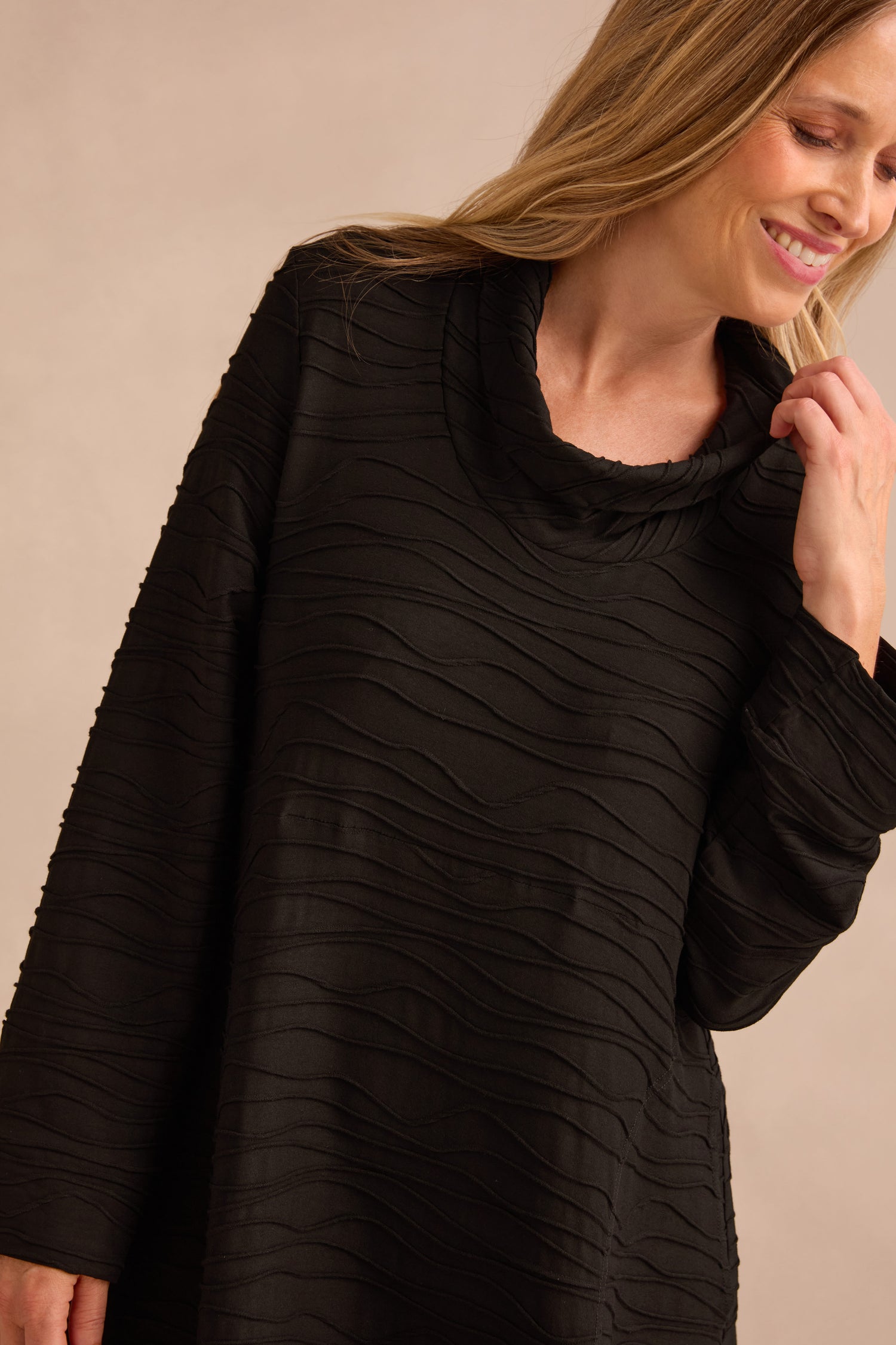 A woman wears the Wave Textured Jersey Cowl Neck Dress in black, featuring long sleeves and a textured finish, as she smiles and looks down against a neutral background.