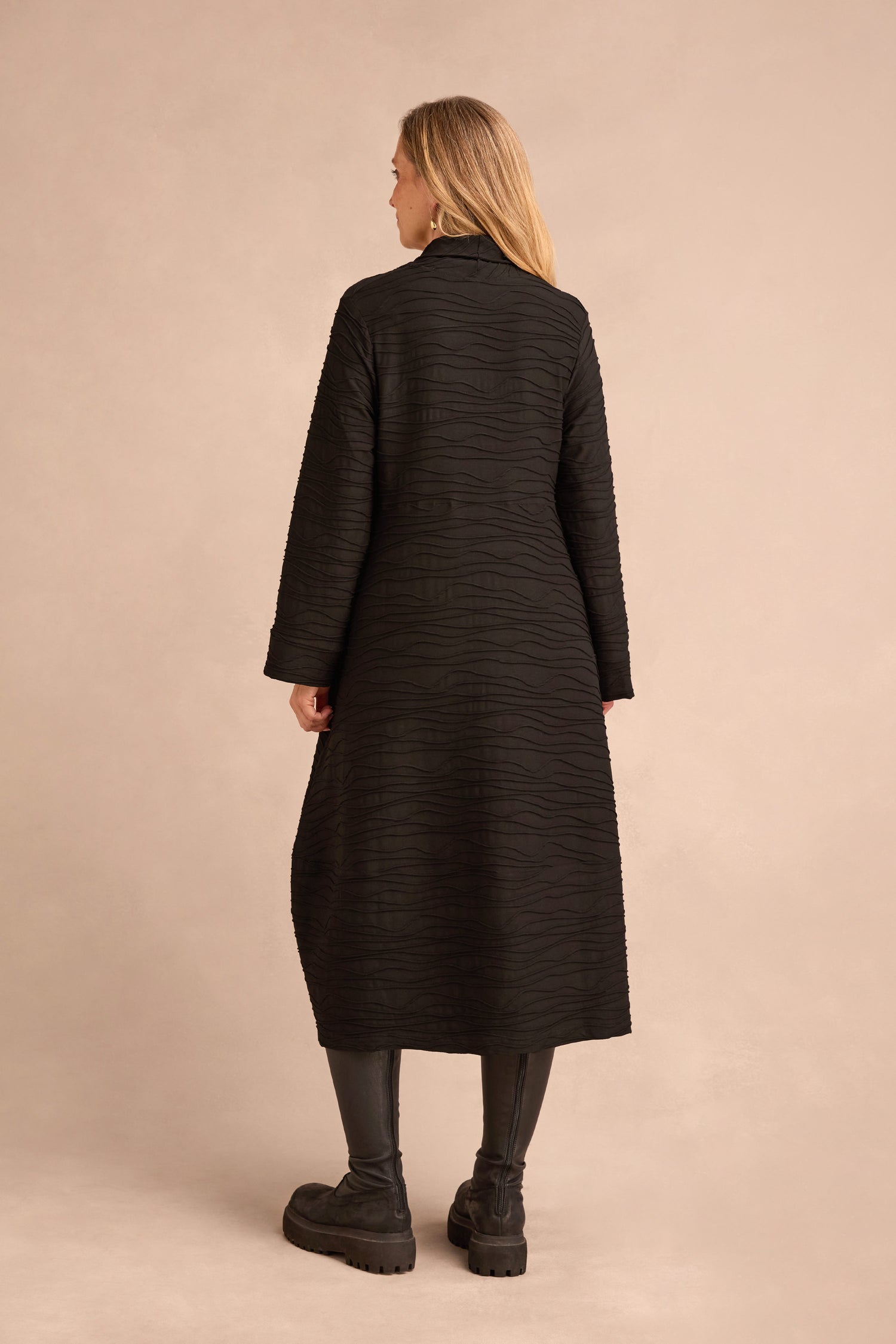 Facing away from the camera, a person wears the Wave Textured Jersey Cowl Neck Dress styled as a coat, paired with black leggings and chunky black shoes against a beige background.