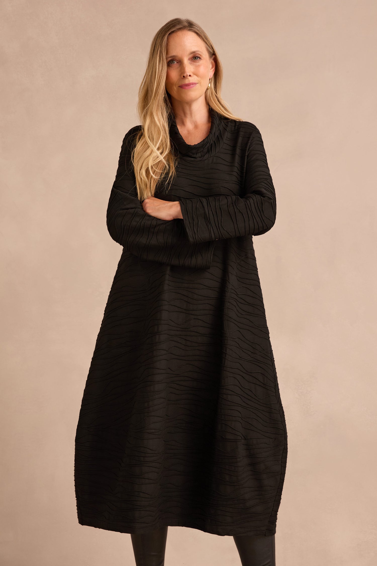 A woman with long blonde hair stands with arms crossed, wearing the Wave Textured Jersey Cowl Neck Dress in black and black leggings, set against a plain beige background.