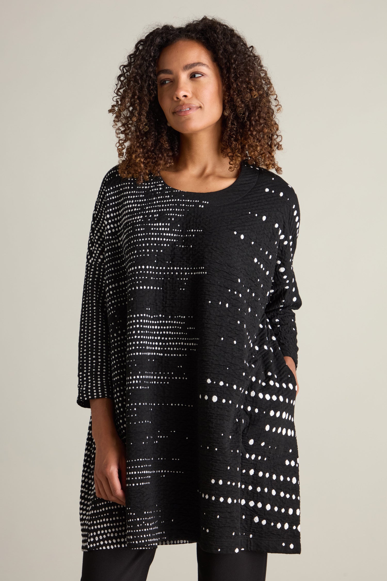 A woman with curly hair is wearing the Sofia Multi Size Spot Jersey Tunic, featuring a relaxed silhouette. Her loose-fitting black tunic, adorned with a white dot pattern, pairs perfectly with her laid-back pose—one hand in her pocket as she gazes slightly off to the side.