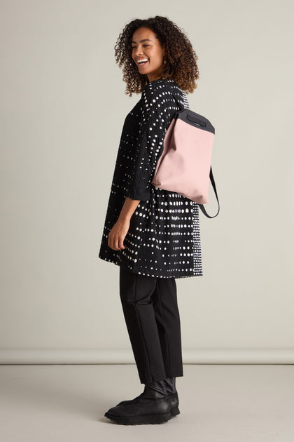 A woman with curly hair, dressed in a Sofia Multi Size Spot Jersey Tunic, black pants, and black boots, stands smiling with a pale pink backpack, perfectly showcasing a relaxed silhouette and eco-friendly fashion.