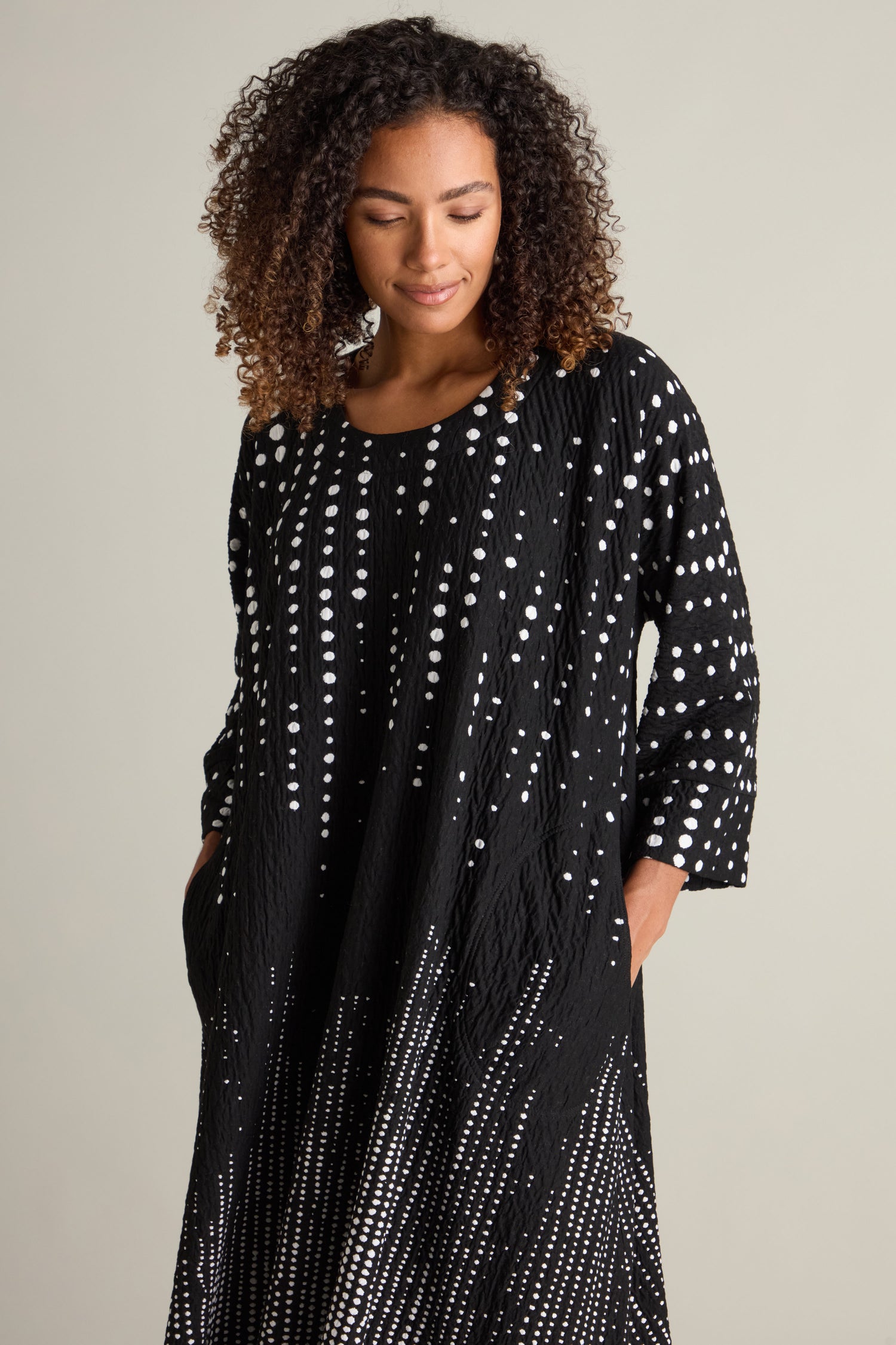 A person with curly hair is wearing a Tiffany Multi Size Spot Jersey Dress made of premium jersey fabric, featuring a white dot pattern. They have their hands in their pockets.