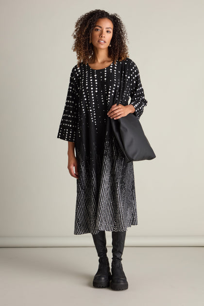 A woman stands against a plain background, wearing a Tiffany Multi Size Spot Jersey Dress with white dots, black boots, and holding a black tote bag—an effortless picture of ecological fashion.