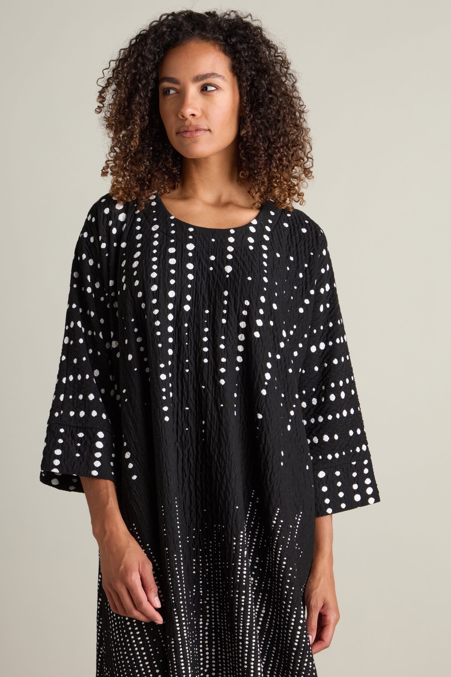 A person with curly hair stands against a plain background, wearing a Tiffany Multi Size Spot Jersey Dress in black with white dot patterns.