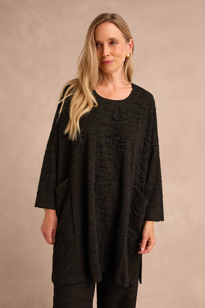 A woman with long blonde hair wears the Textured Squares Jersey Tunic, featuring a loose fit, long sleeves, pockets, and a textured design, while standing against a neutral beige background.