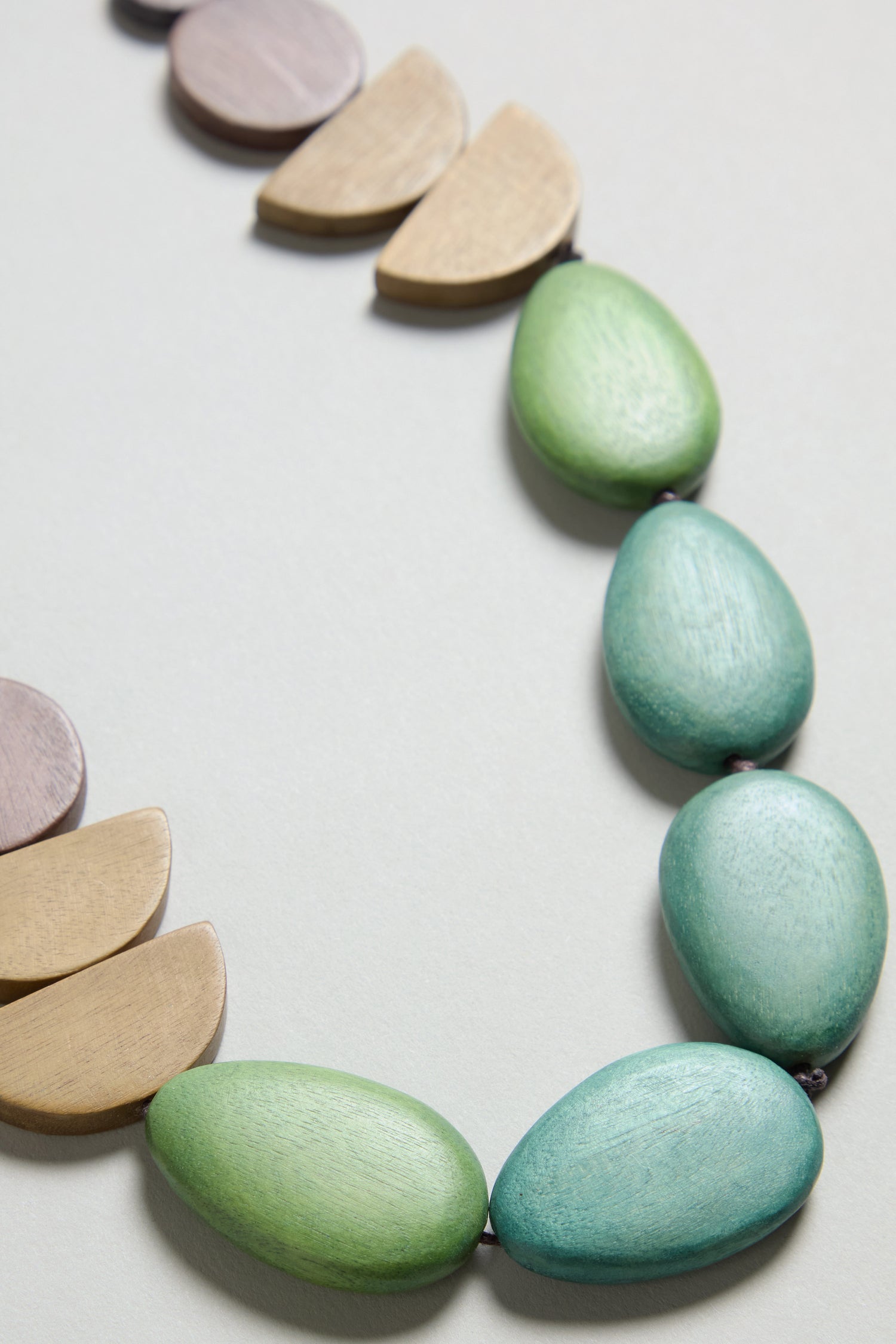 Close-up of the Assorted Wooden Shapes Necklace: an adjustable cord featuring oval green and teal beads with half-moon beige and mauve beads, displayed on a light surface.