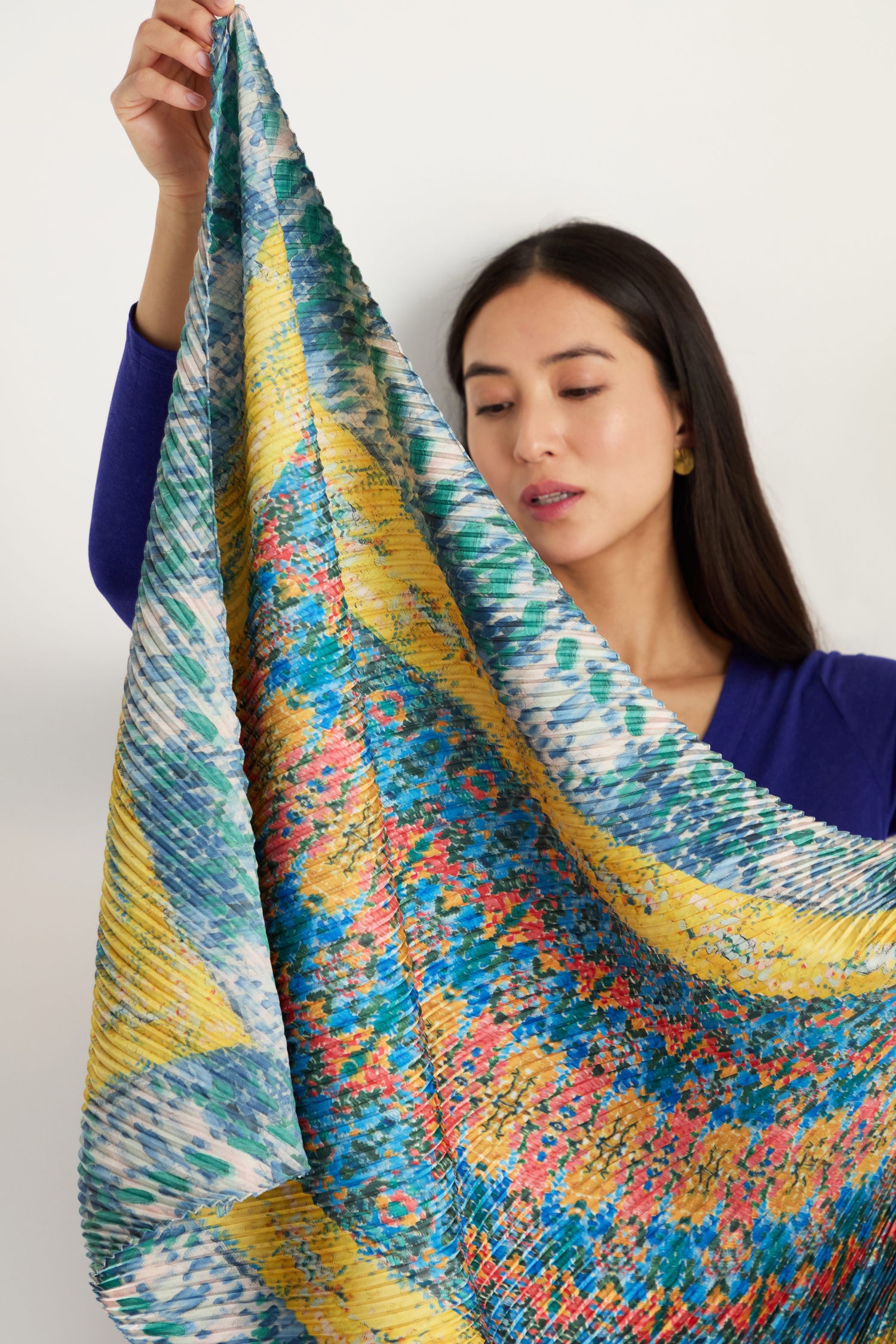 A woman in a blue top holds up the Artistry Pleat Scarf, a large, colorful pleated satin accessory with blue, yellow, and red designs—lightweight and perfect for versatile styling.