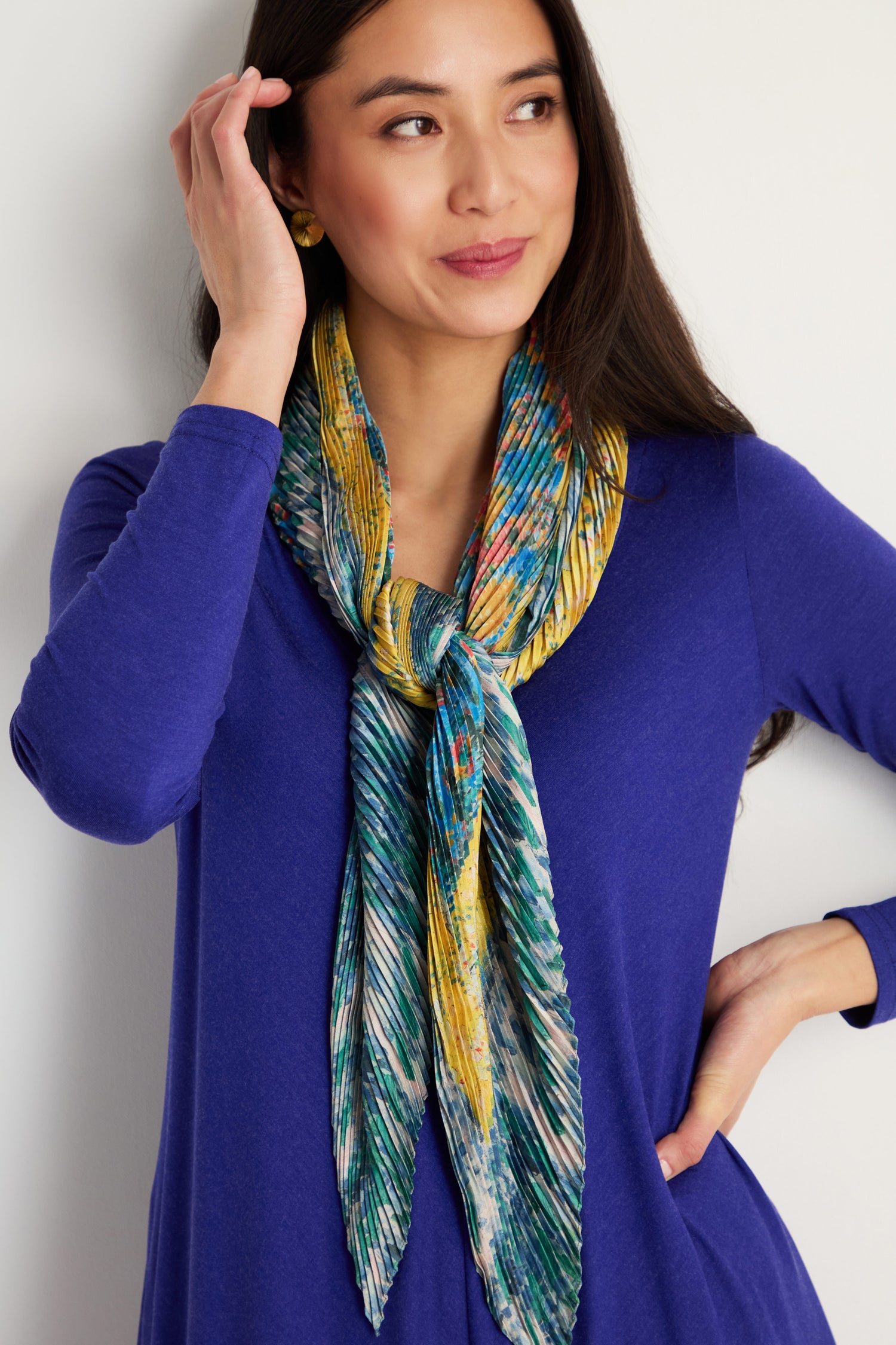 Woman wearing a blue long-sleeve top styles the Artistry Pleat Scarf—a colorful, lightweight statement accessory—against a light background, with one hand in her hair and the other on her hip for versatile flair.