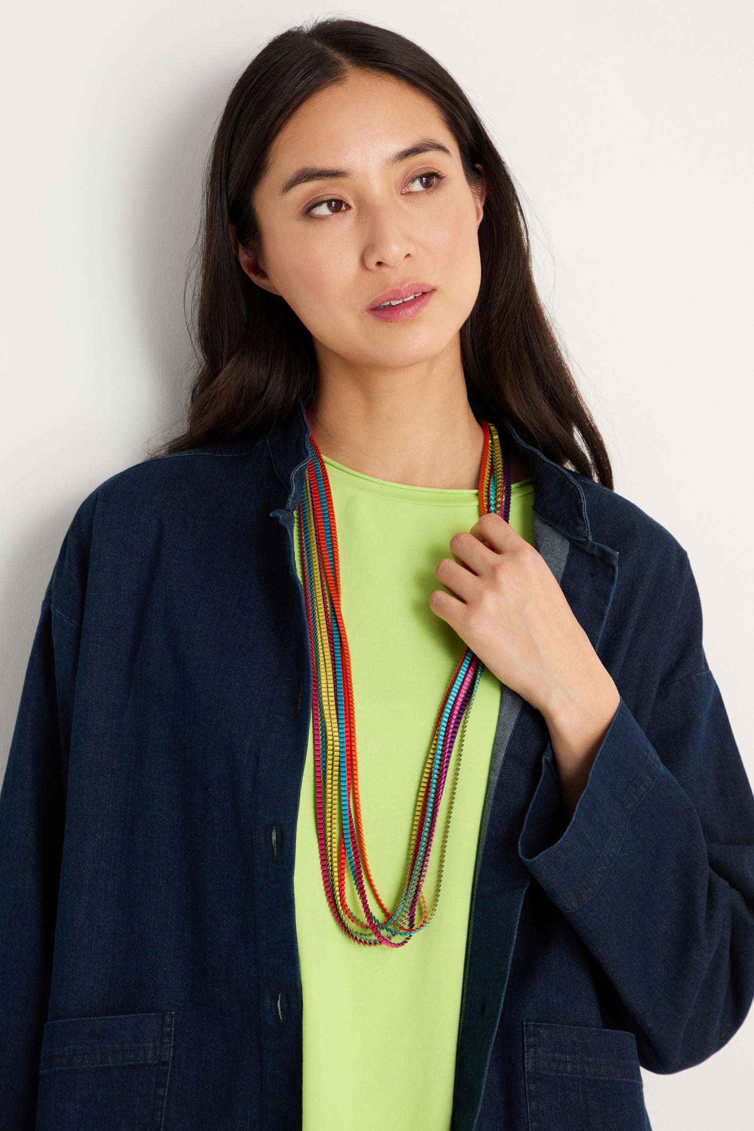 A woman with long dark hair stands against a plain background, wearing a denim jacket over a light green shirt and showcasing the Long Pleated Necklace.