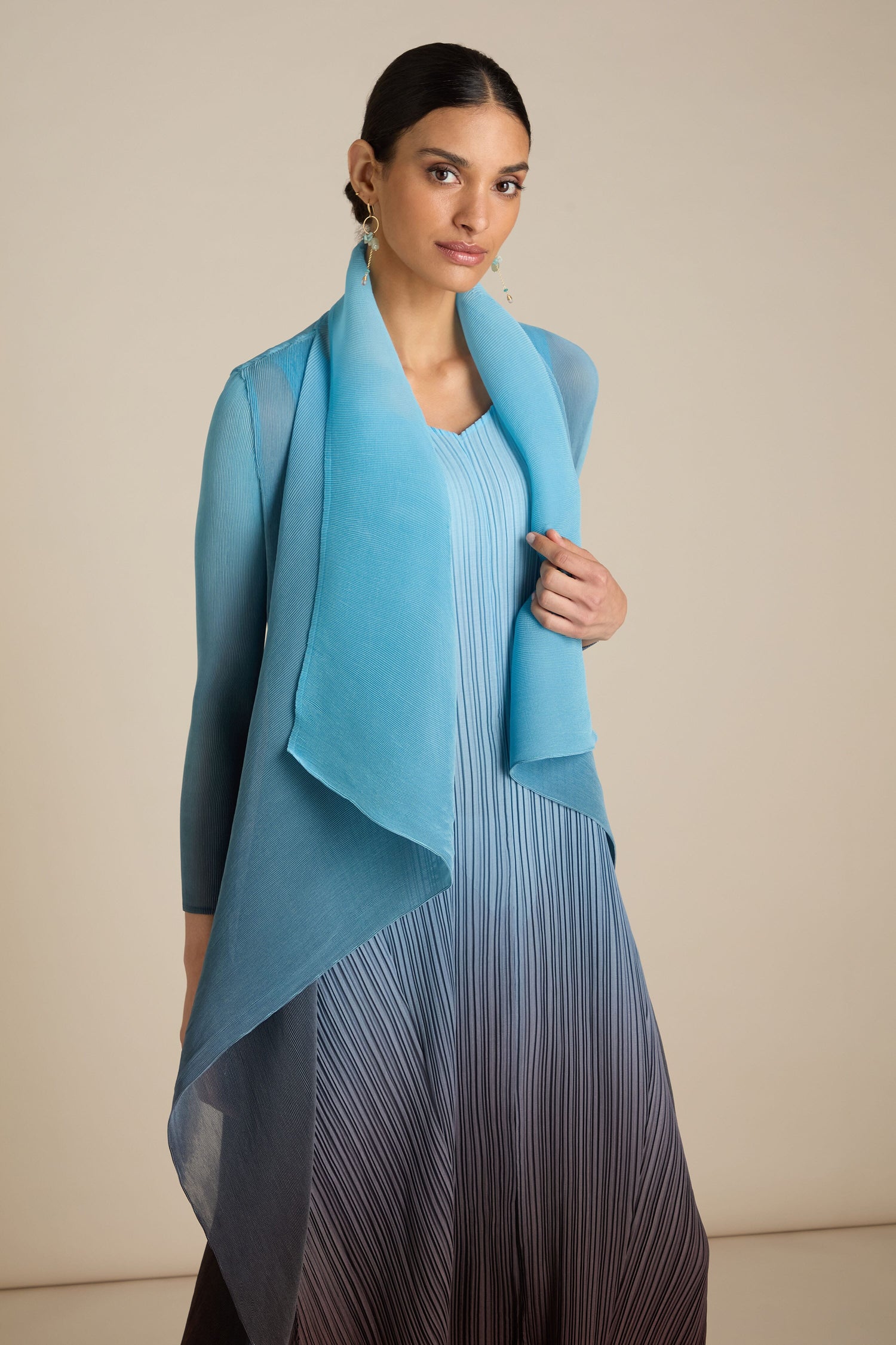 A person stands posing in a studio, wearing a layered blue dress paired with a long, flowing, semi-transparent blue scarf and complemented by the Pleated Jacket.