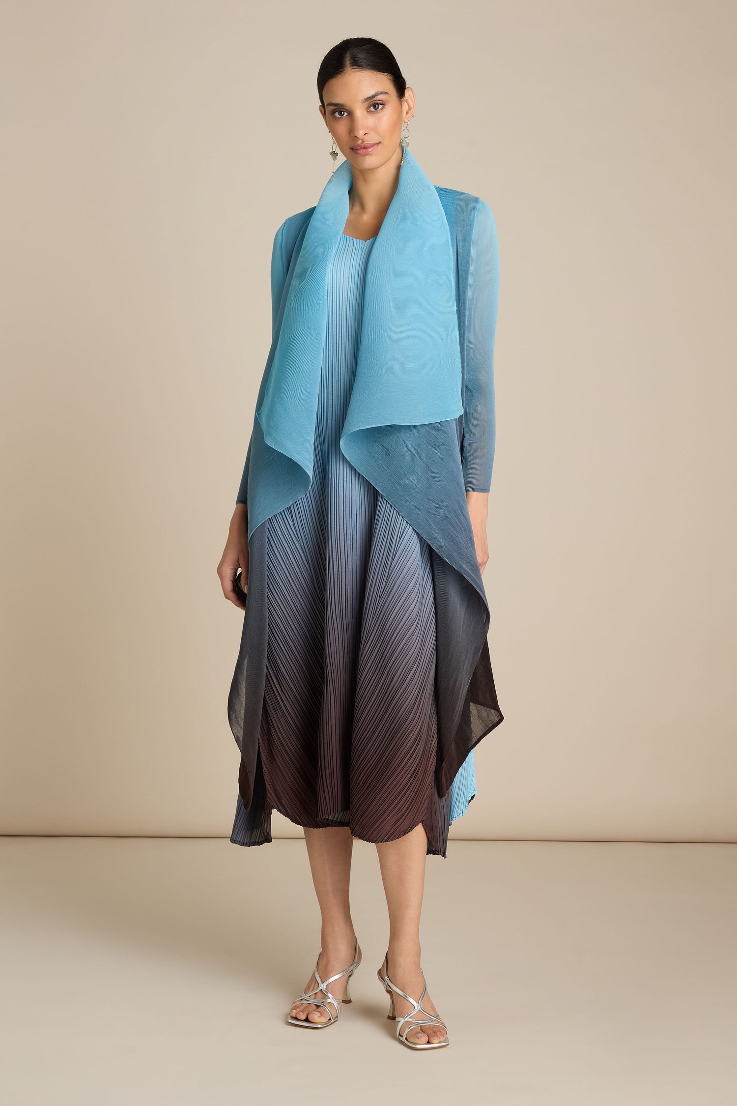 A woman stands wearing a flowing, gradient dress with hues of blue, transitioning to a darker shade towards the hem, paired with silver sandals. She dons the Pleated Jacket—a handcrafted piece that showcases exquisite Japanese craft—in front of a plain beige background.