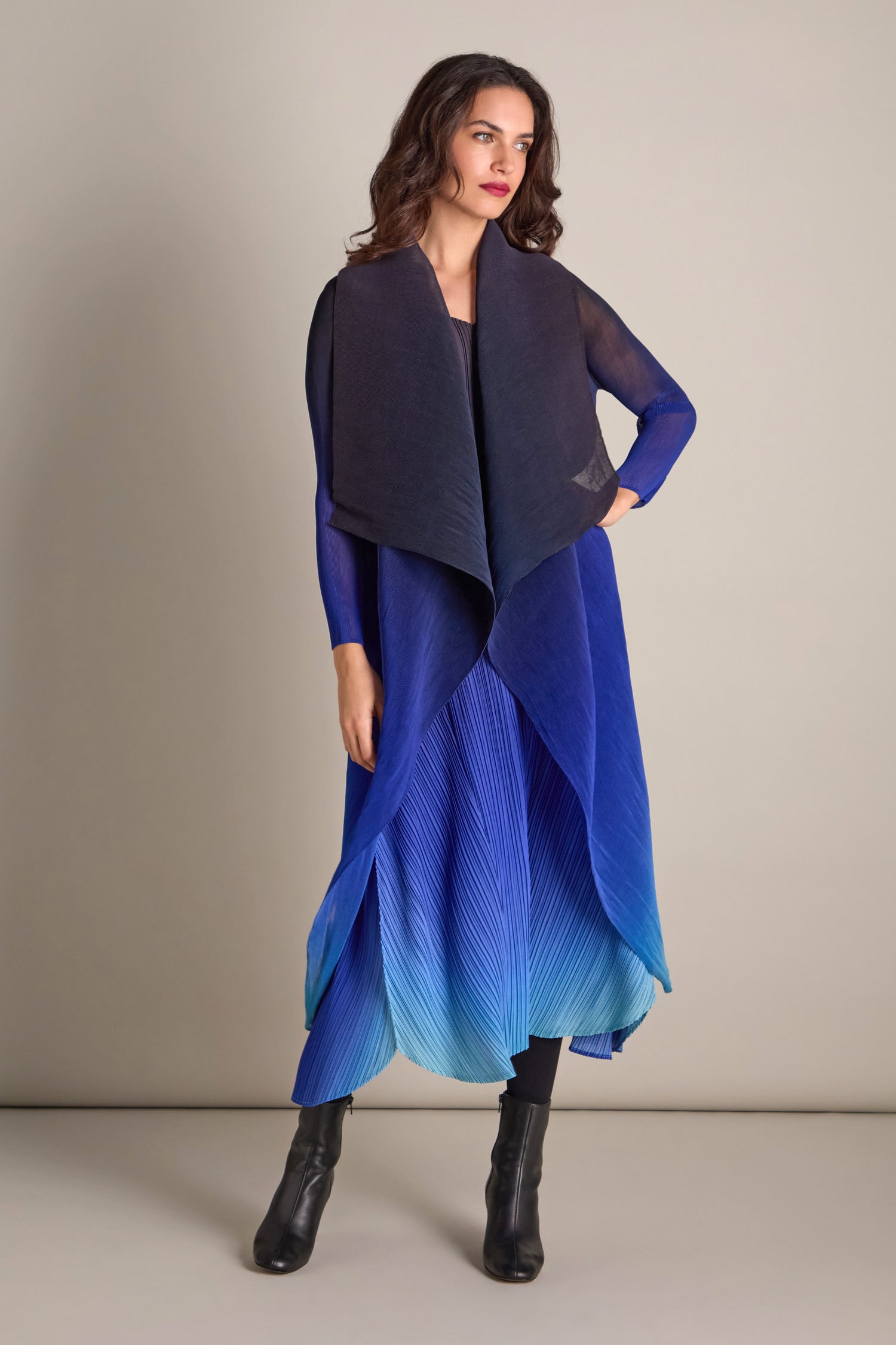 A person stands against a plain backdrop wearing a handcrafted, flowing gradient blue dress paired with black boots and a sheer black shawl, complemented by the Pleated Jacket.