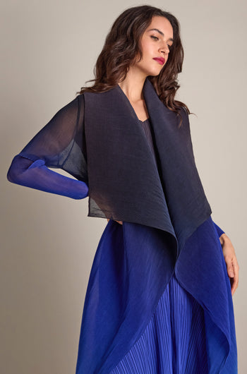 A woman with long hair wears a flowing Pleated Jacket in blue and black, featuring a Japanese shibori pattern, as she poses against a neutral background.