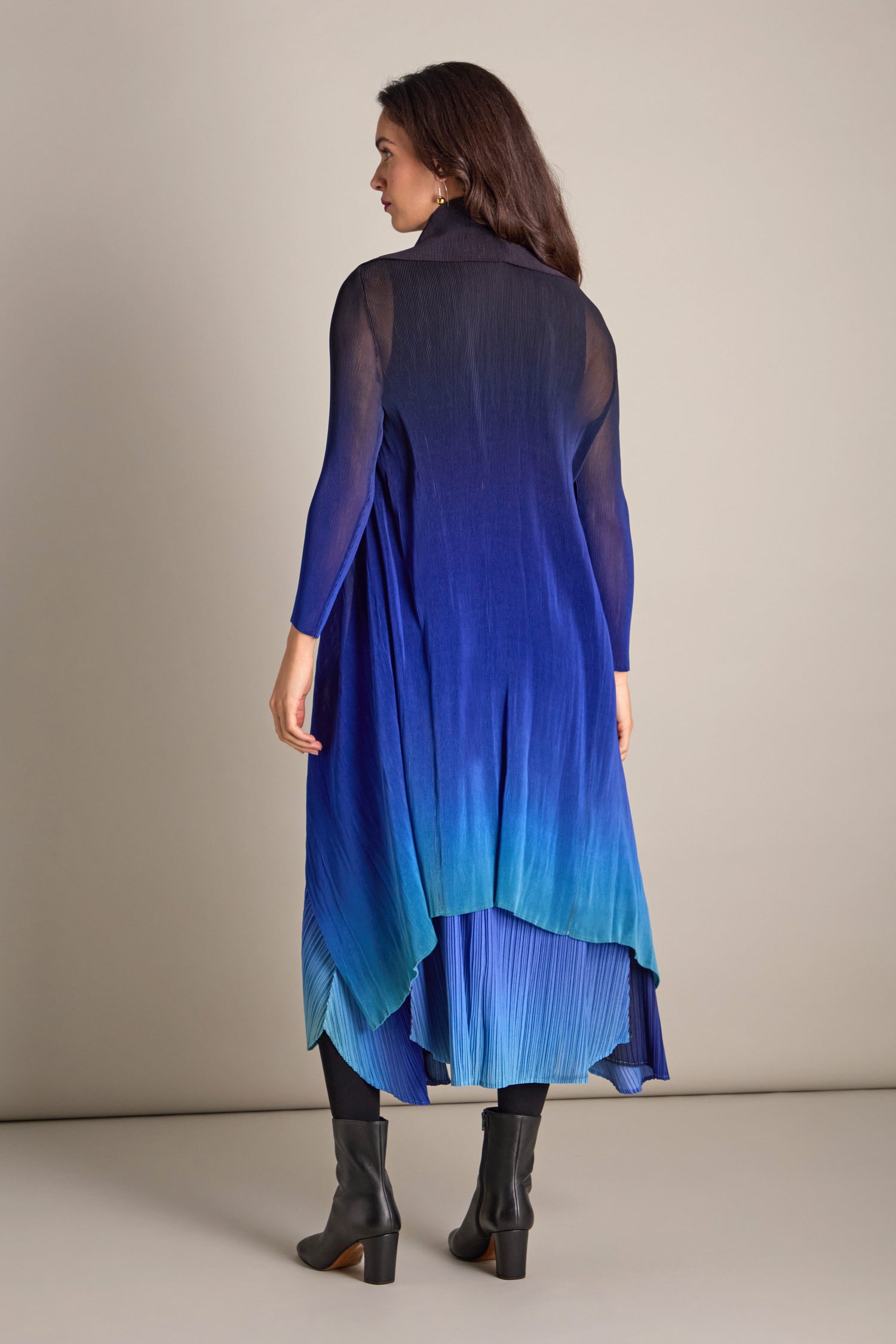 A woman in a blue ombre dress featuring a pleated skirt and black boots is seen from behind in a neutral studio setting. The addition of the Pleated Jacket adds elegance, beautifully complementing the Japanese shibori-inspired design on her attire.