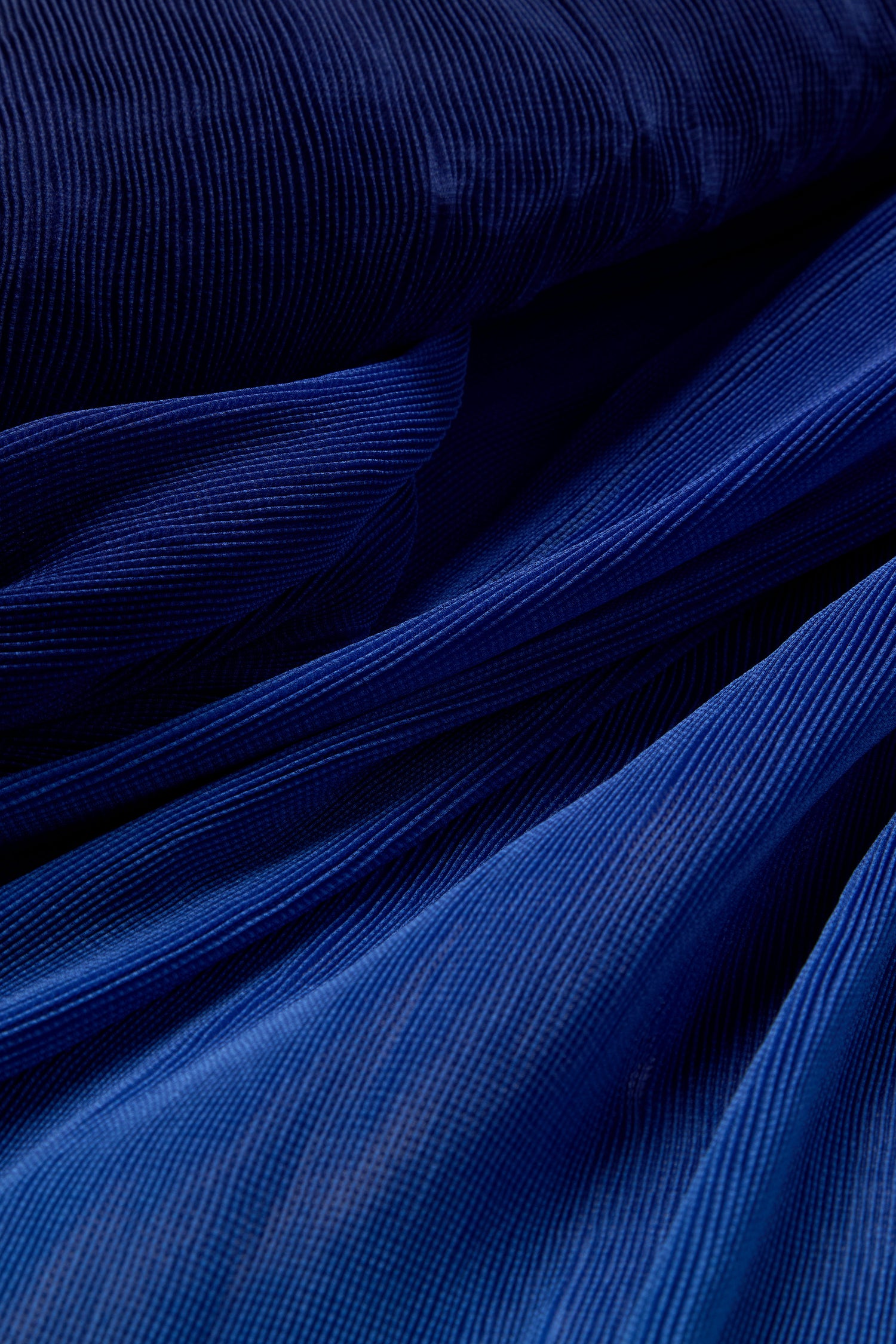 Close-up of a Pleated Jacket in flowing dark blue fabric with fine vertical stripes, reminiscent of Japanese shibori techniques, creating a textured appearance.