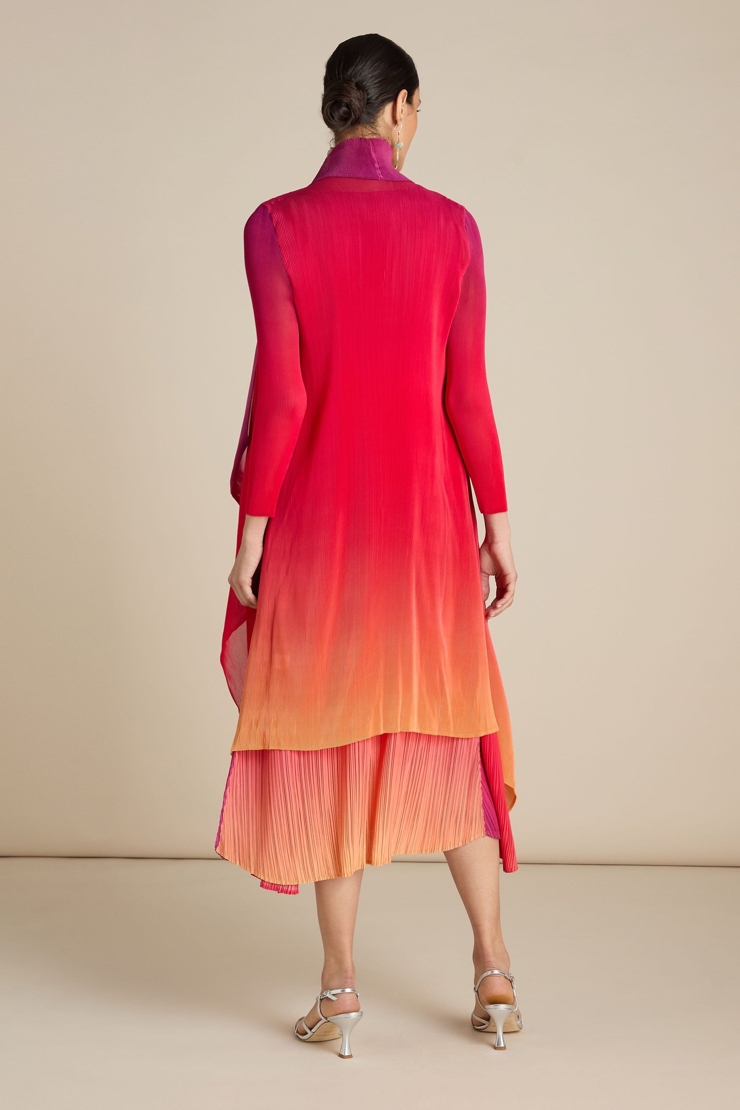 A person stands with their back to the camera, wearing a long-sleeved gradient dress in shades of red, pink, and orange, paired with light-colored open-toed heels. Their hair is styled in a low bun as they drape a Pleated Jacket over one shoulder.