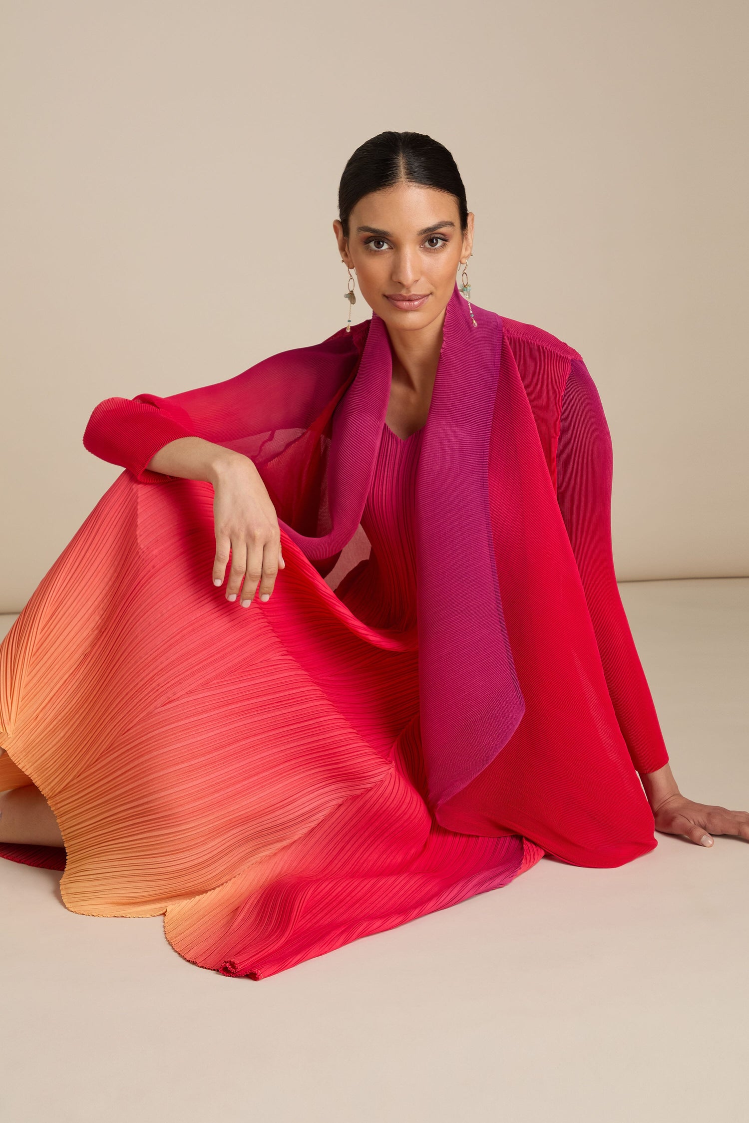 A seated individual in a gradient Pleated Jacket, transitioning from pink to orange, poses against a plain beige background. The person is accessorized with dangling earrings that evoke the elegance of Japanese craft.