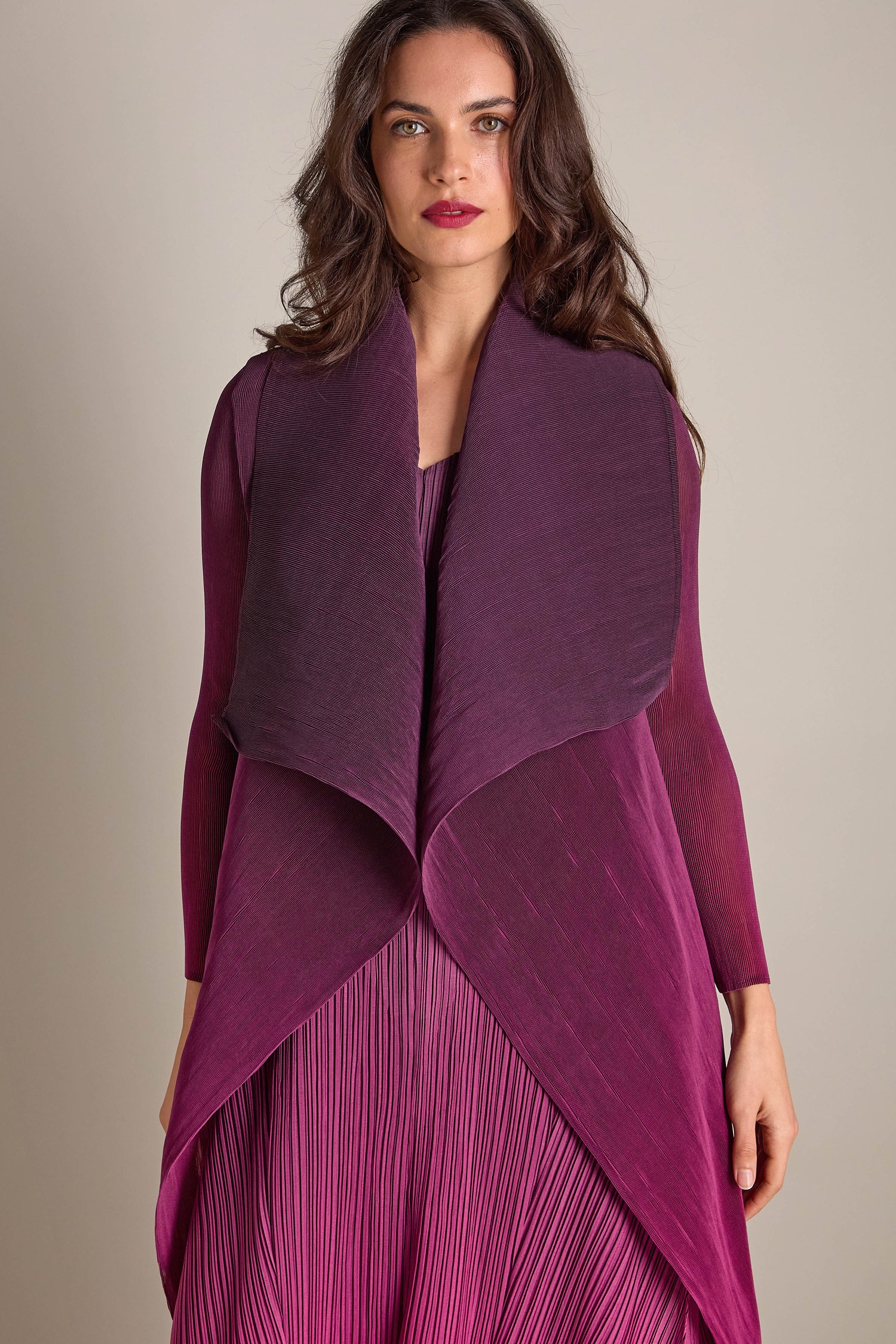 Against a plain backdrop, a person stands elegantly in a pleated magenta jacket paired with a shibori dark shawl.