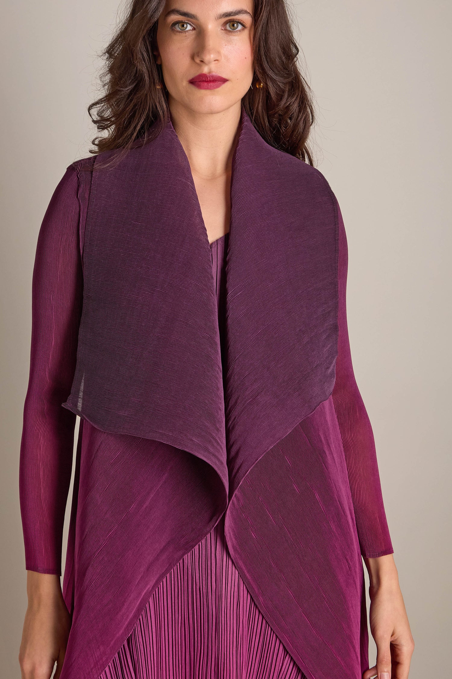 A woman wearing a long, pleated purple jacket over a matching dress stands against a neutral background.