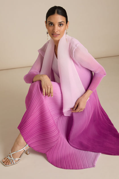 A person sits on a beige background, wearing a gradient pink pleated jacket and silver strappy heels, looking at the camera. They have their hair tied back and wear long drop earrings that showcase exquisite Japanese craft.