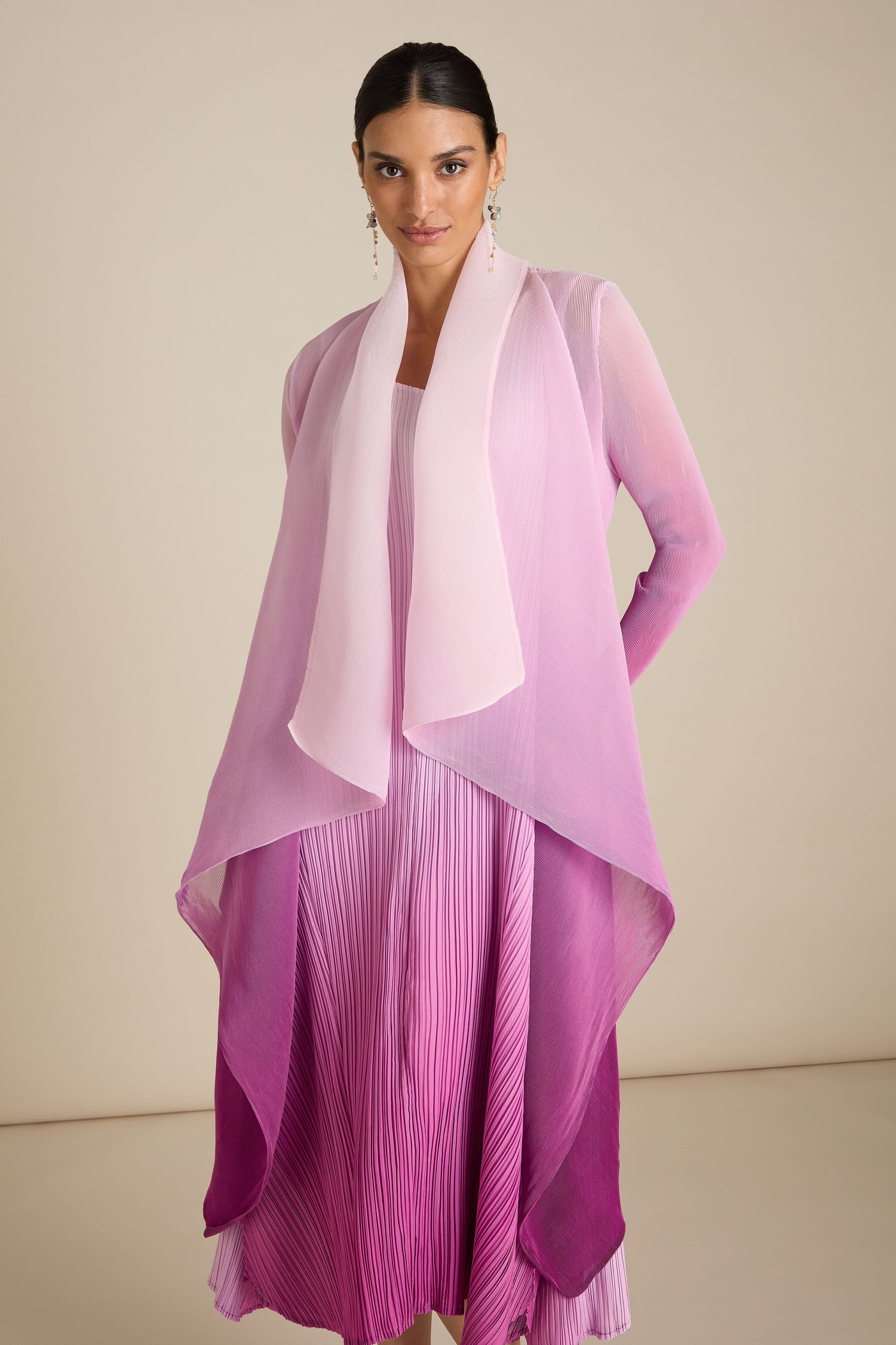 A person stands wearing a pink and purple gradient Pleated Jacket with a matching handcrafted layered shawl. The background is neutral-toned.