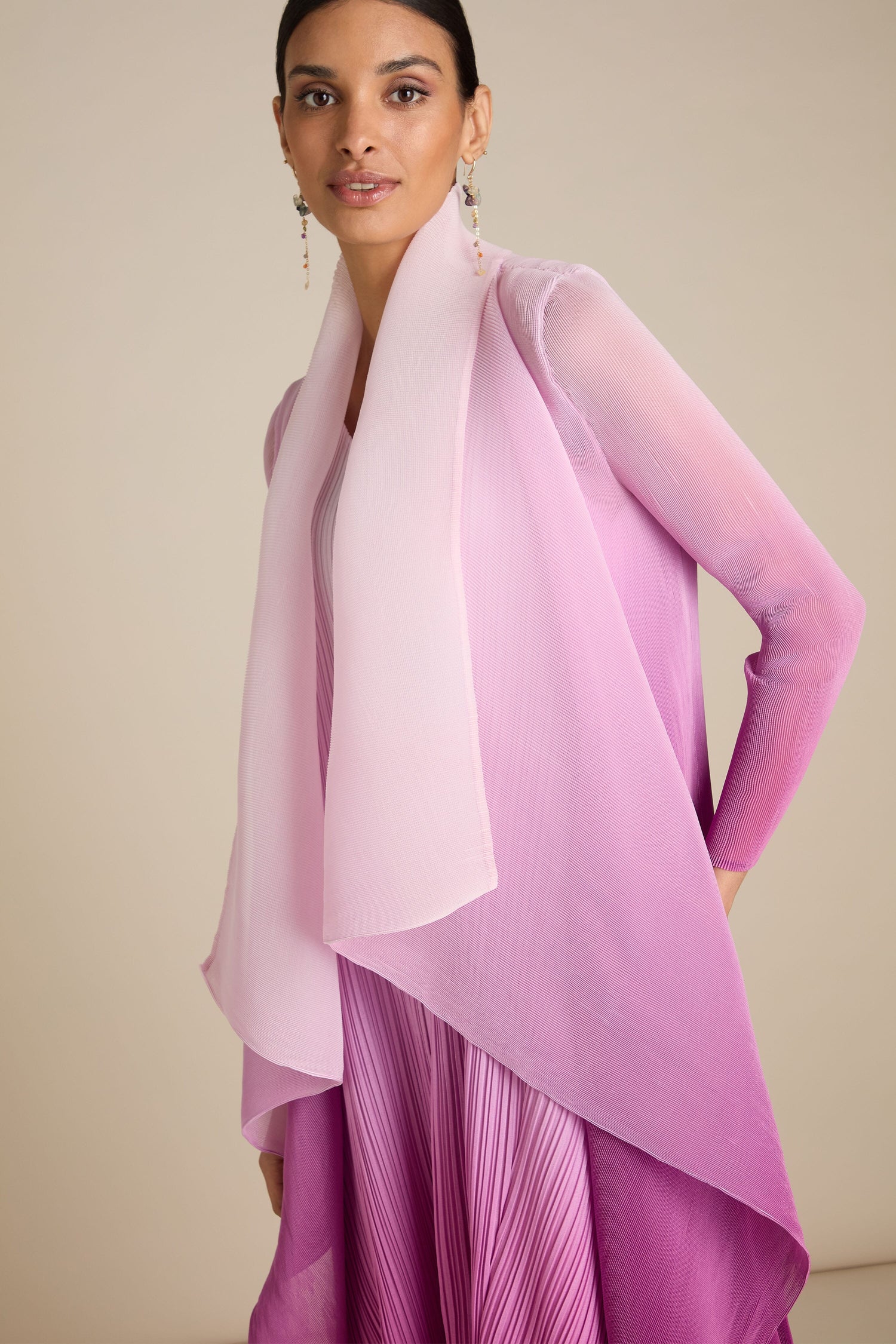 A person stands wearing a pink and purple gradient dress with matching sheer shawl and dangling earrings, complemented by the Pleated Jacket, a handcrafted piece inspired by Japanese craft.