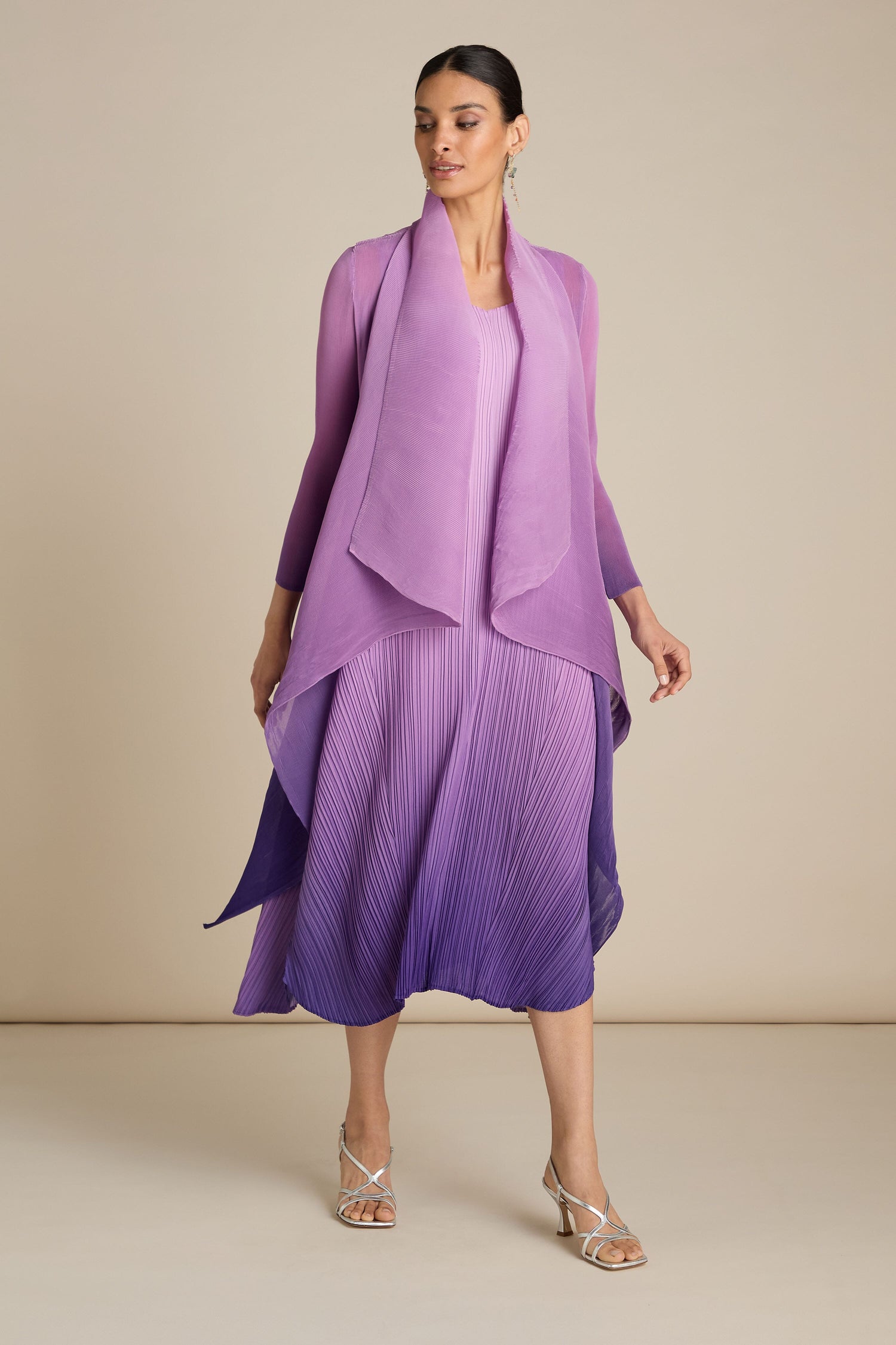 Person stands wearing an elegant, sleeveless, gradient purple Pleated Jacket with a matching shawl. This jacket, reminiscent of Japanese craft, transitions from light lavender at the top to deep purple at the bottom.