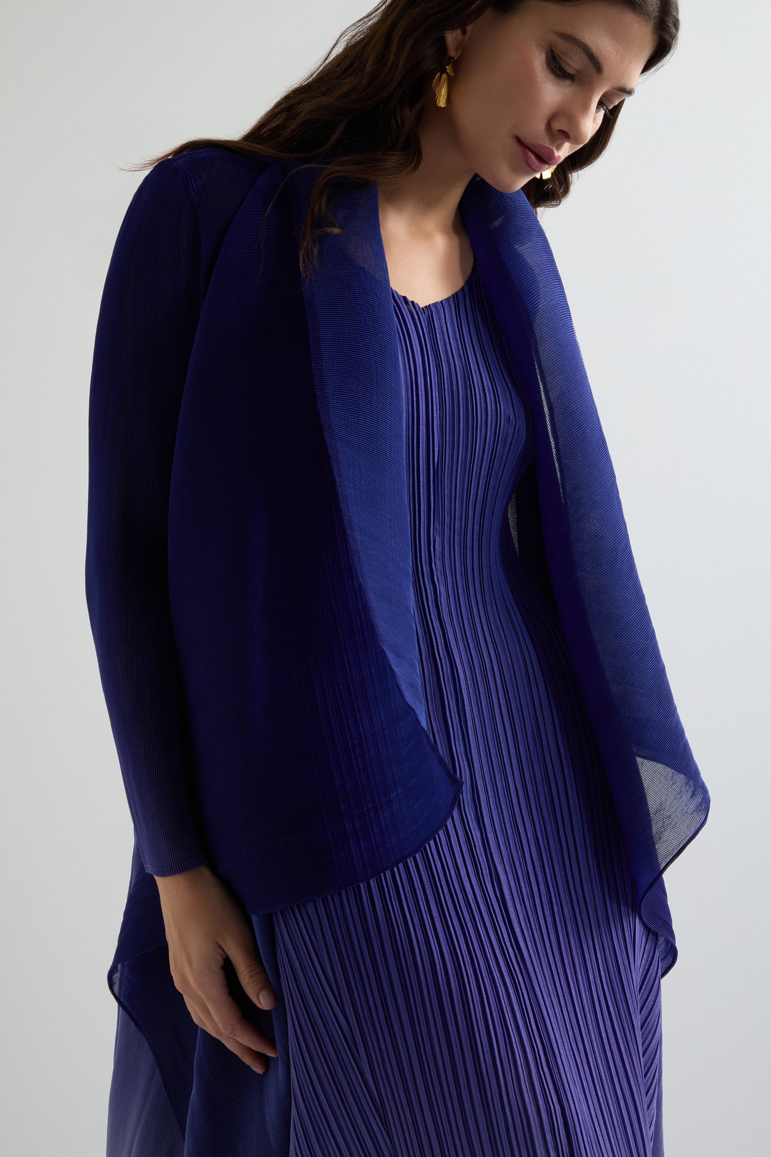 A woman exudes contemporary style in a pleated, deep blue dress paired with a matching sheer pleated jacket, radiating elegance as she looks downward.