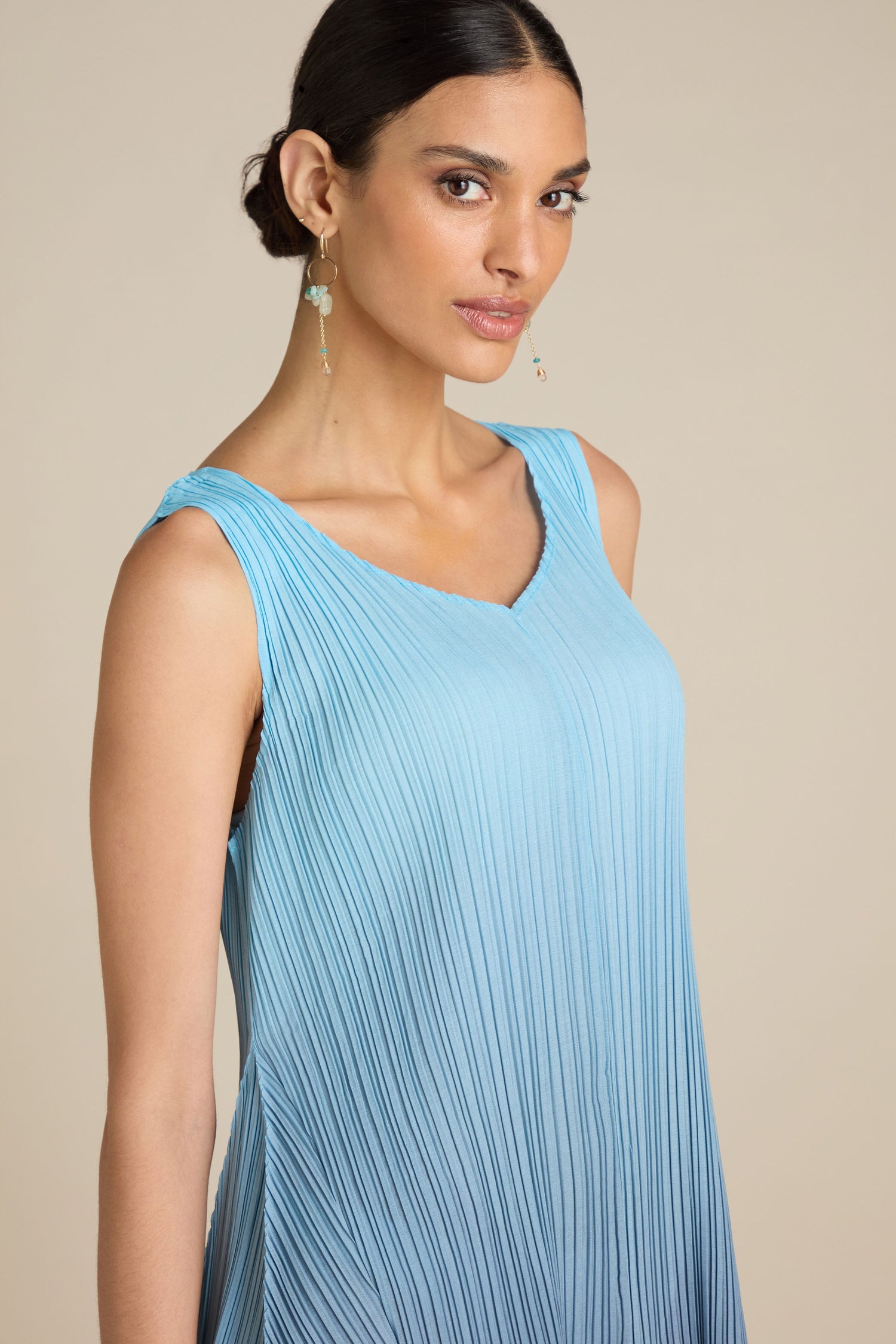 A woman with her hair pulled back wears a light blue Pleated Dress with a sleeveless, relaxed silhouette, paired with dangling earrings, while looking towards the camera with a neutral expression.