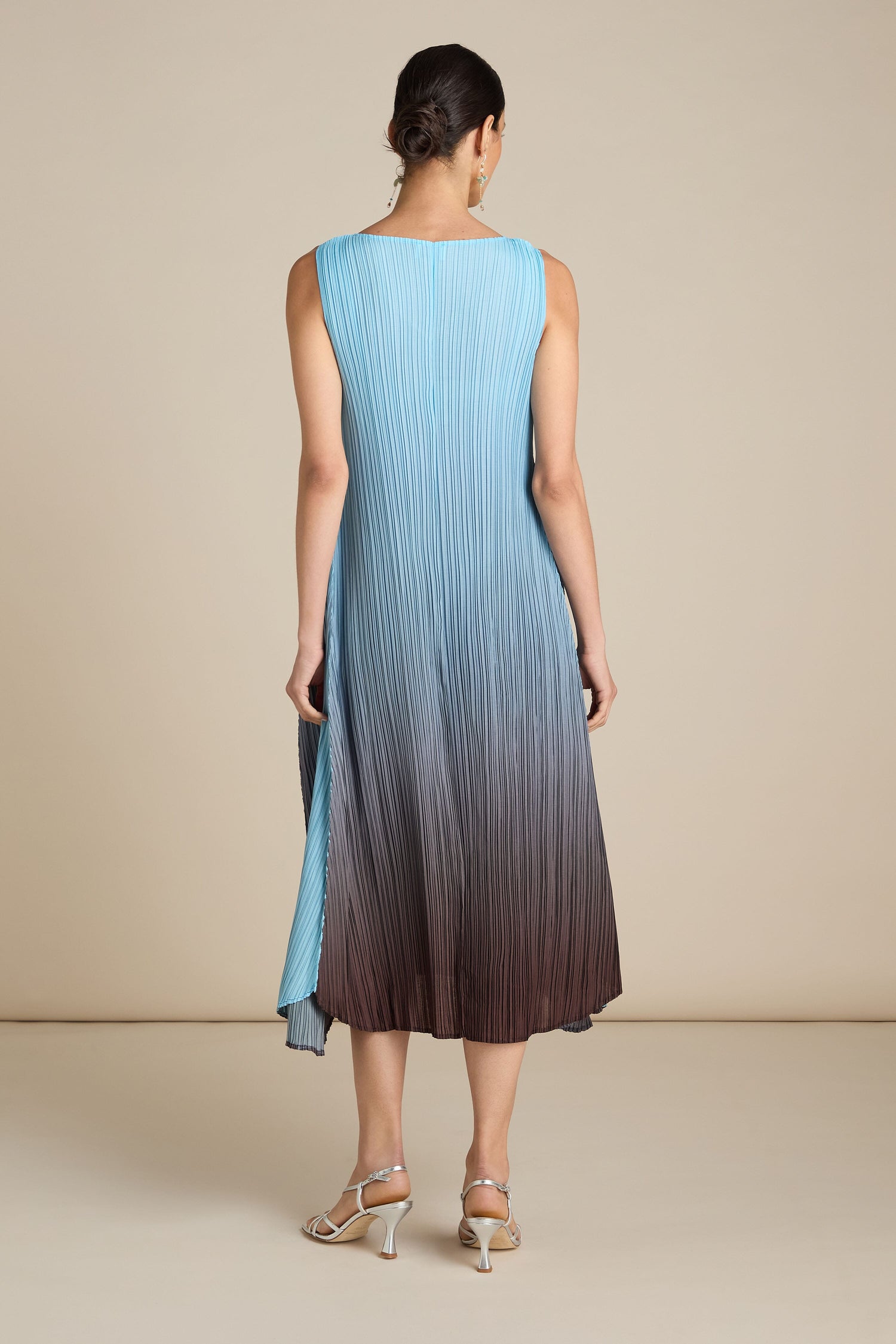 A woman with her back to the camera wears a Pleated Dress featuring a relaxed silhouette, which transitions from light blue at the top to dark brown at the bottom. She completes her look with strappy sandals, adding a touch of bohemian flair.