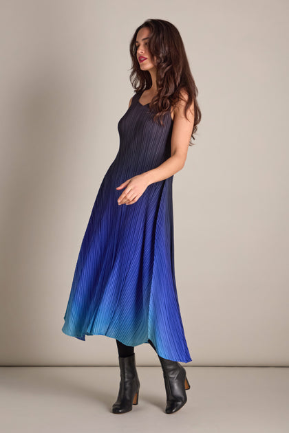 A person stands against a plain backdrop wearing a sleeveless Pleated Dress in gradient blue, featuring intricate pleated fabric, paired with black ankle boots.