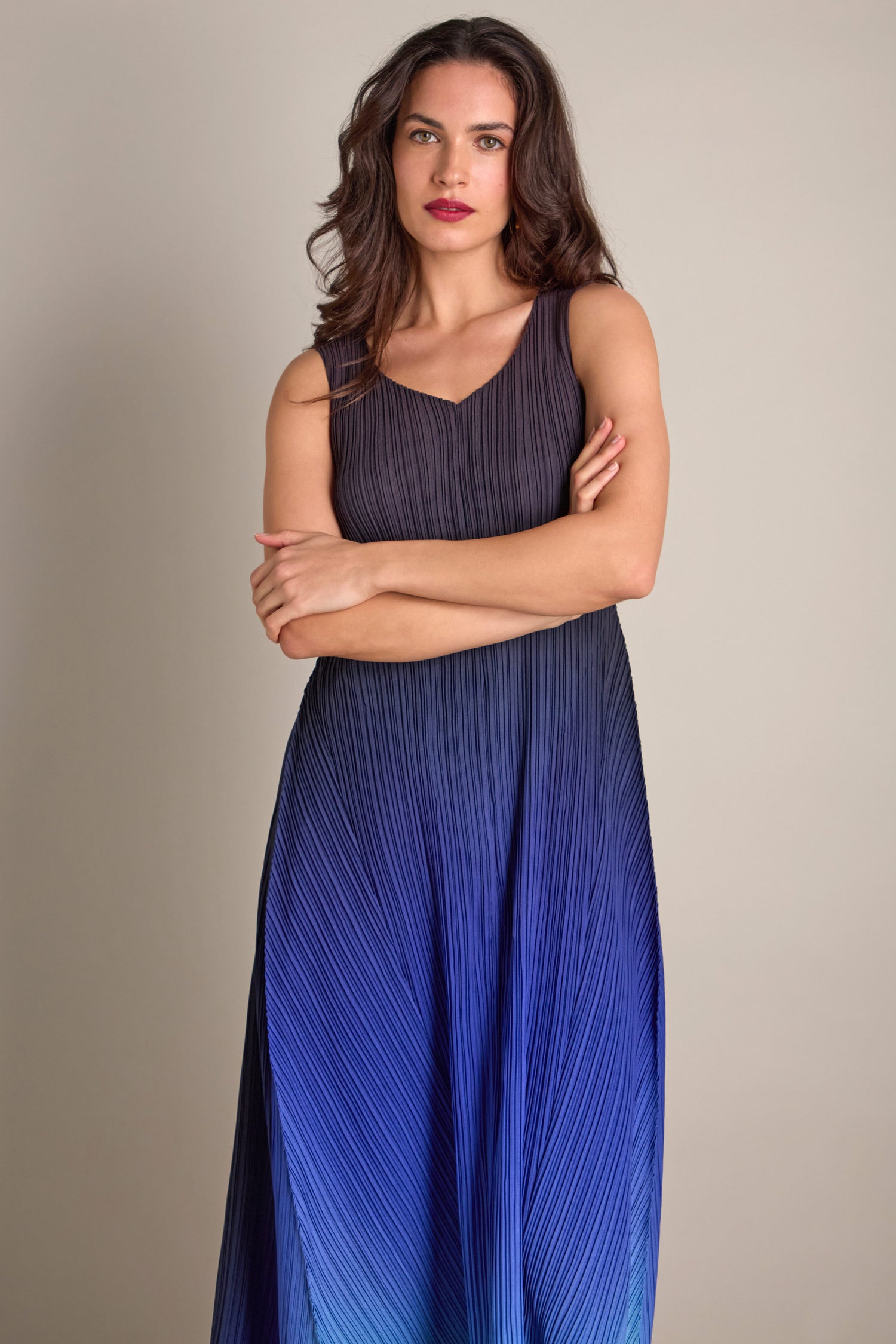 A person stands with arms crossed, dressed in a sleeveless Pleated Dress showcasing a gradient of blue hues and intricate pleats, offering a bohemian touch.