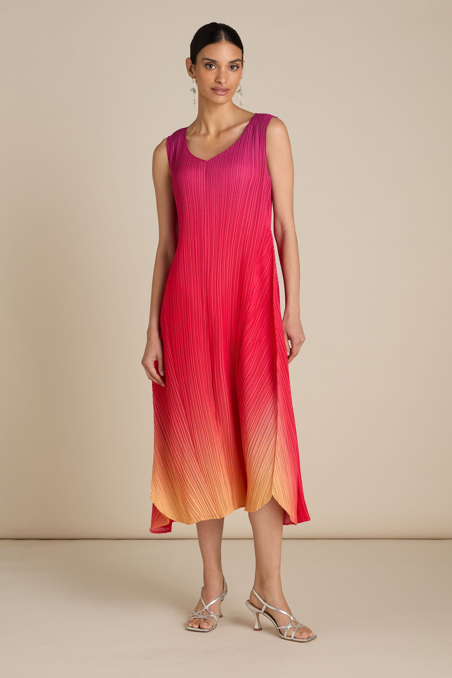 A person stands against a plain background wearing the Pleated Dress, featuring a sleeveless design that transitions from pink at the top to orange at the bottom. They have paired it with metallic sandals. The relaxed silhouette of the dress adds a touch of bohemian flair to the ensemble.