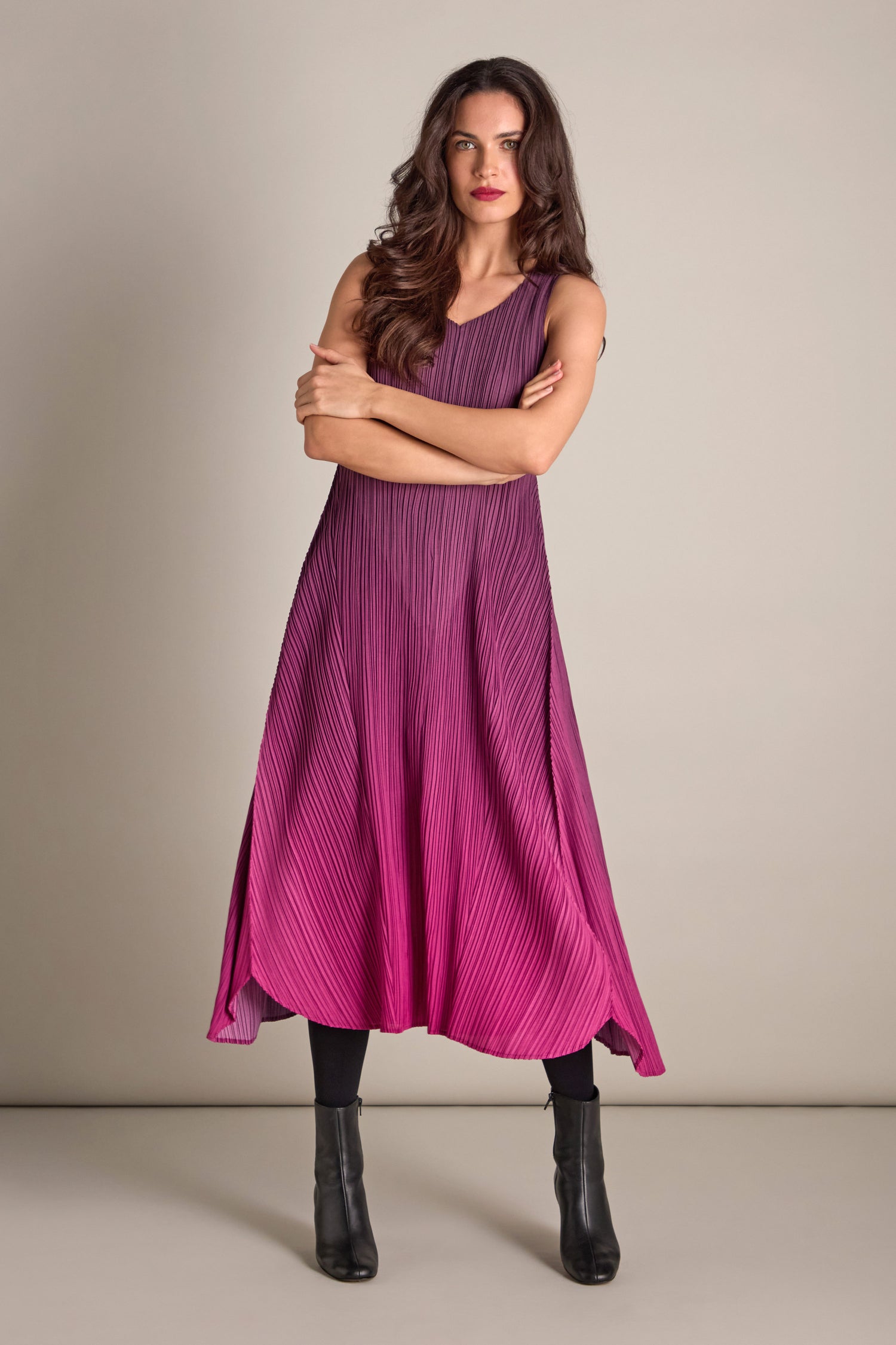 Wearing a bohemian style pleated dress paired with black boots, a person with long hair stands confidently against a plain background, their arms stylishly crossed.