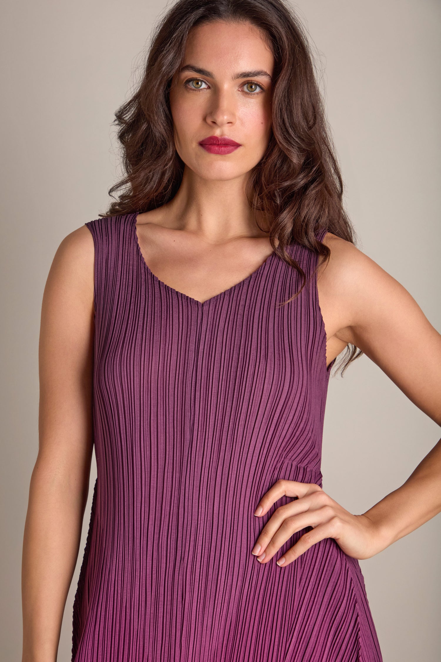 A woman with long brown hair wears a sleeveless Pleated Dress in a rich purple shade, its pleated fabric adding a bohemian touch as she stands against a neutral backdrop.