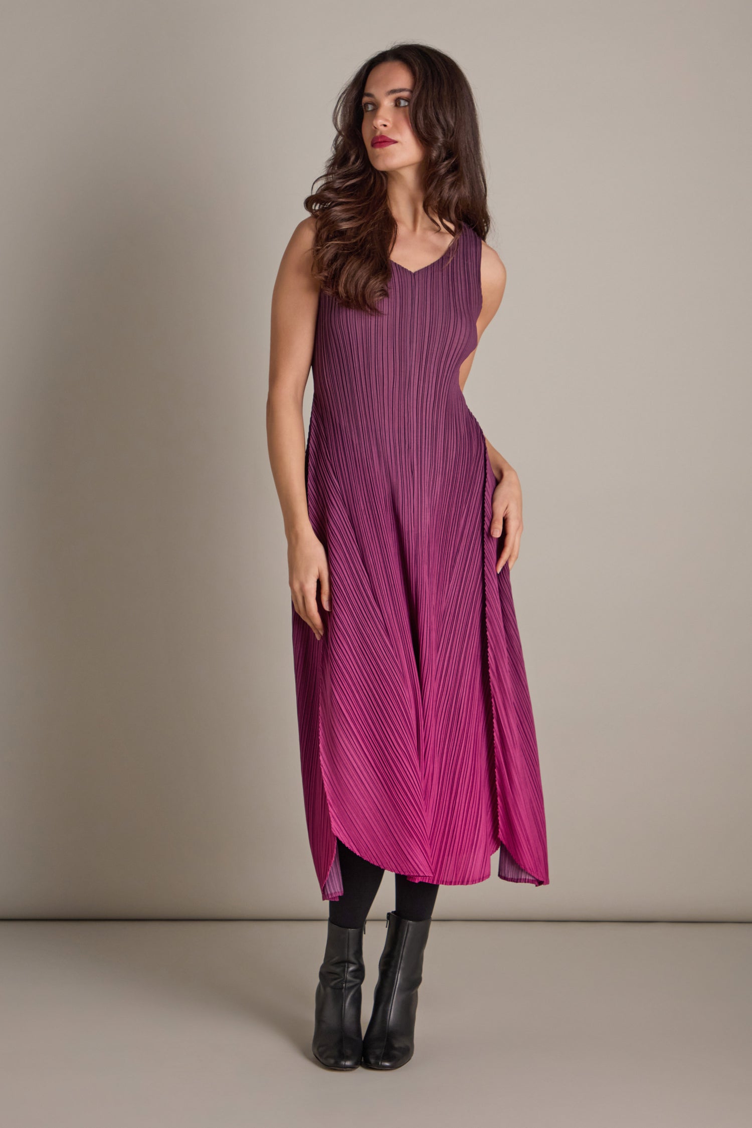 A woman in a sleeveless, pleated dress in plum stands against a neutral backdrop. She showcases bohemian flair with her chic black ankle boots.