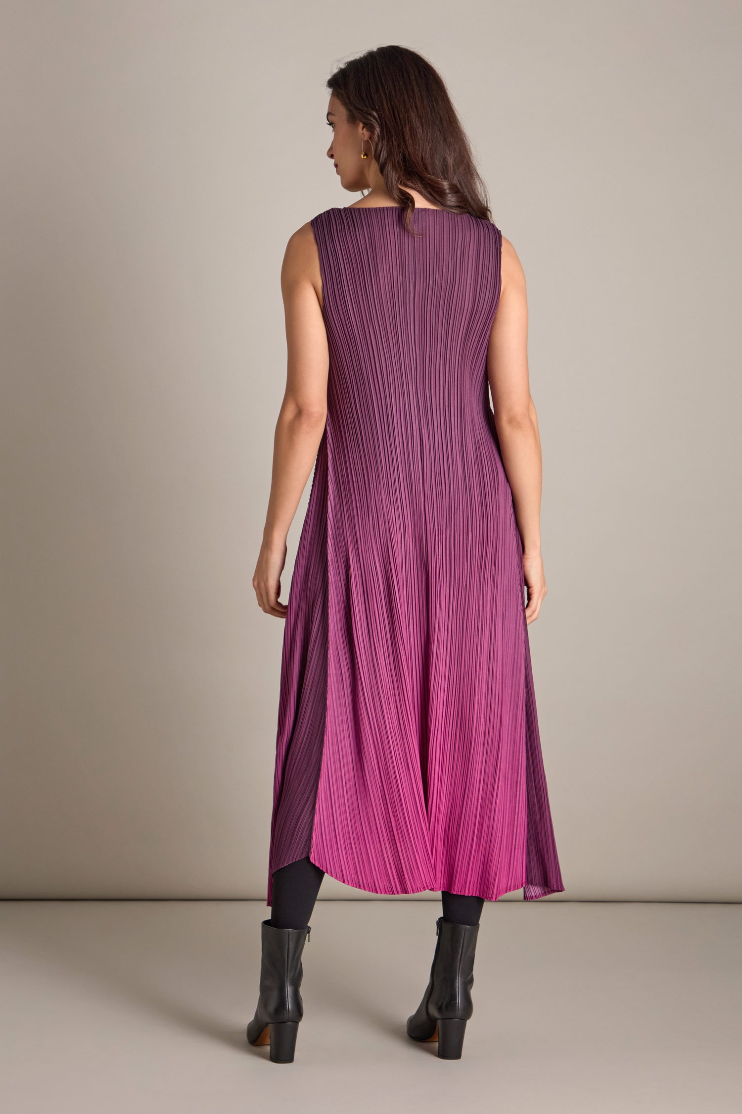 A woman stands wearing the Pleated Dress in gradient shades of purple and pink, paired with black boots, exuding a subtle bohemian flair as she faces away.