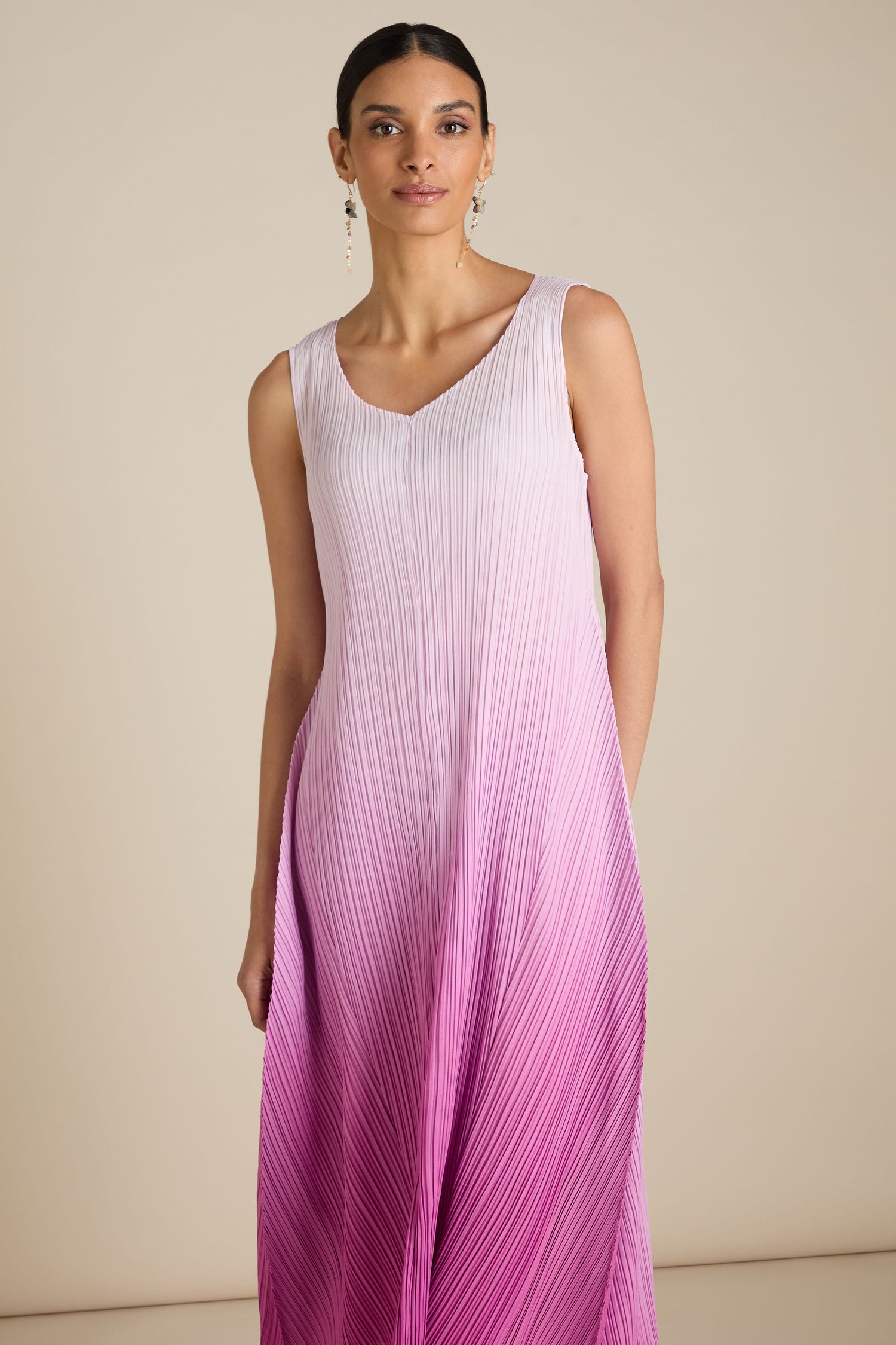 A model wearing the Pleated Dress, which seamlessly transitions from white at the top to pink at the bottom, stands against a plain background. The dress showcases a relaxed silhouette infused with a touch of bohemian elegance.